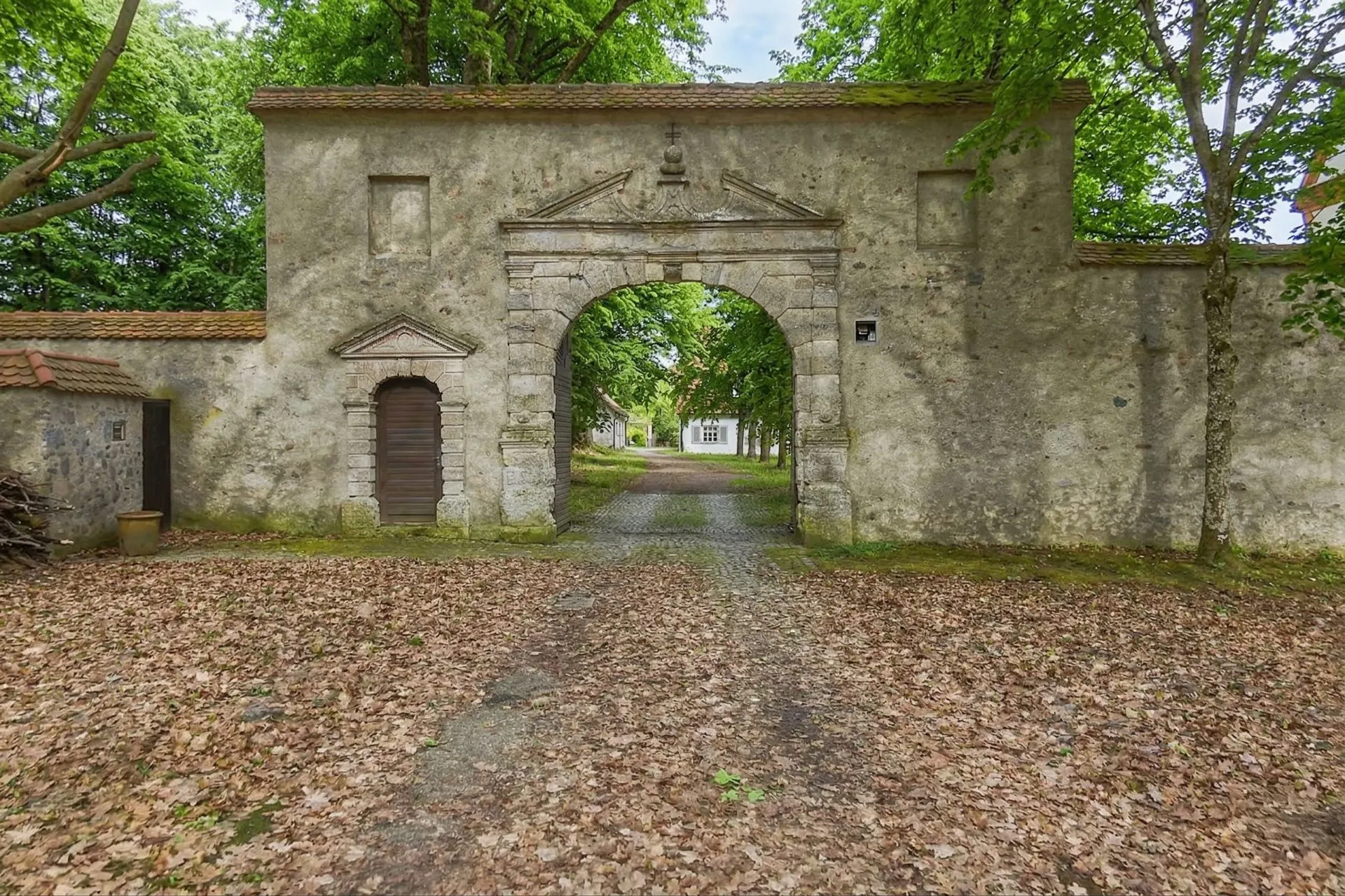 Altes steinernes Tor mit Rundbogen und Holztür, von Bäumen umgeben.