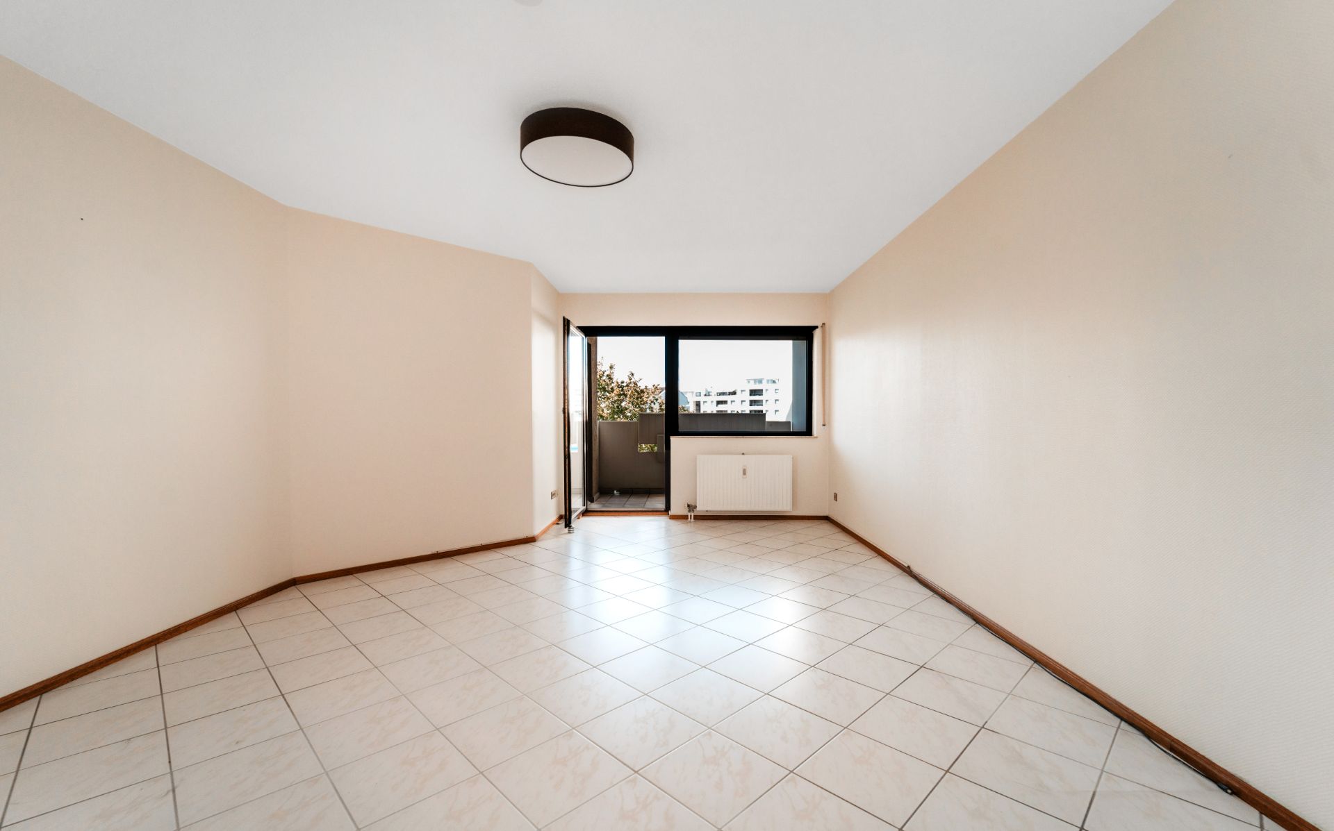 Leerer Wohnraum mit weißen Fliesen, großem Fenster und Zugang zu einem kleinen Balkon.