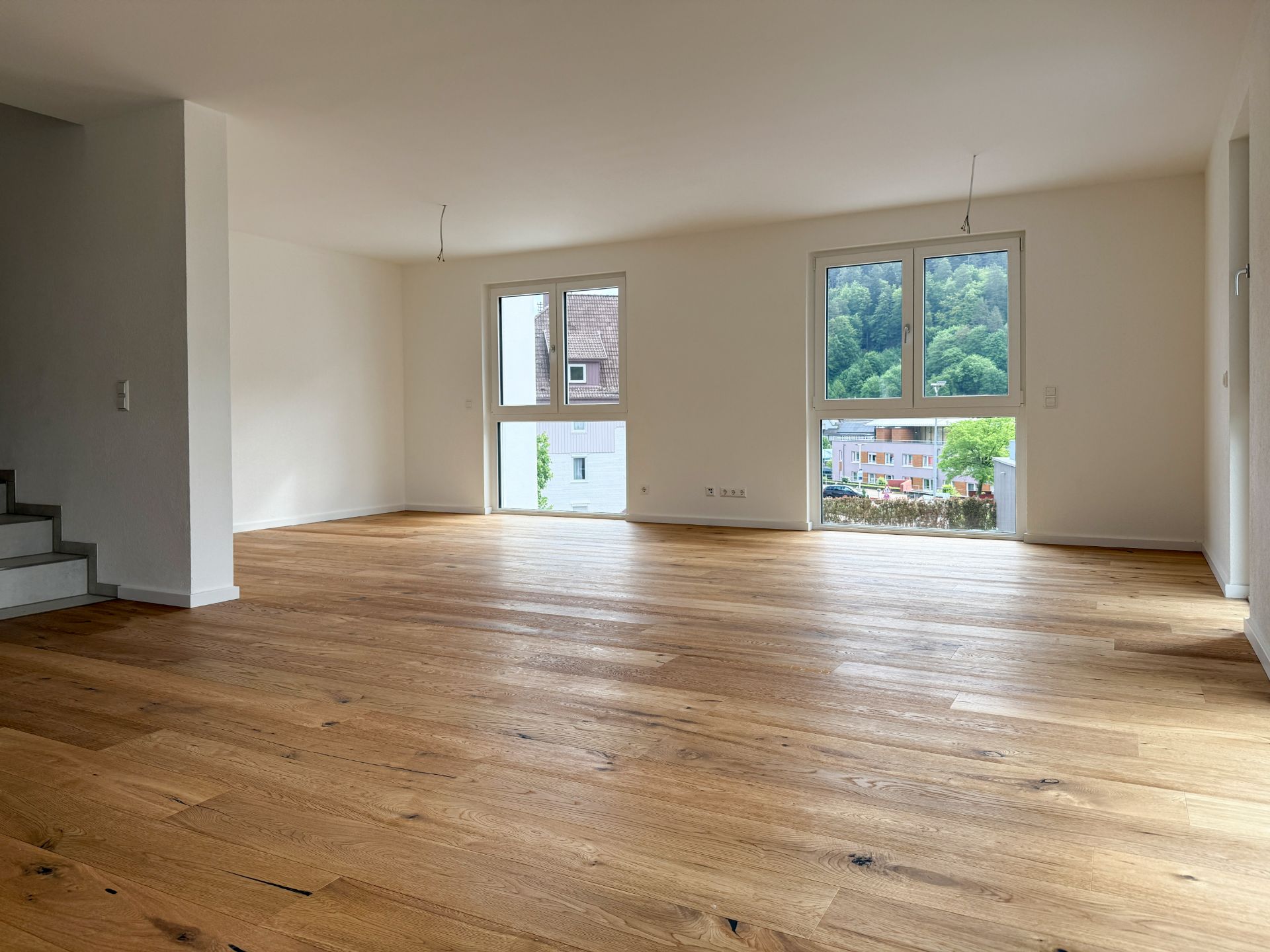Helles, leeres Zimmer mit Holzfußboden und großen Fenstern, Blick auf Bäume und Häuser.