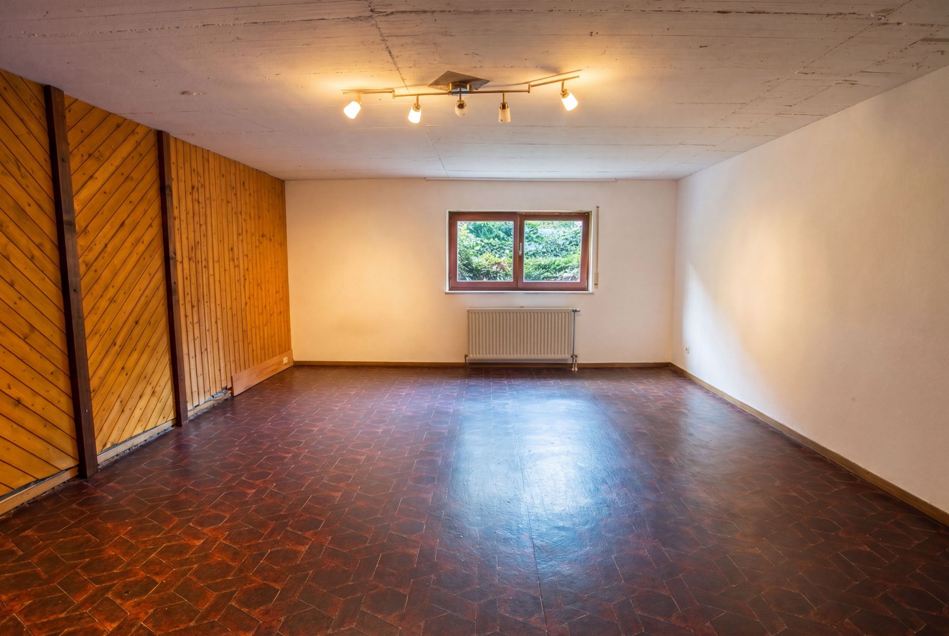 Leerer Raum mit dunklem Boden, Holzvertäfelung an einer Wand, Fenster und Heizkörper.