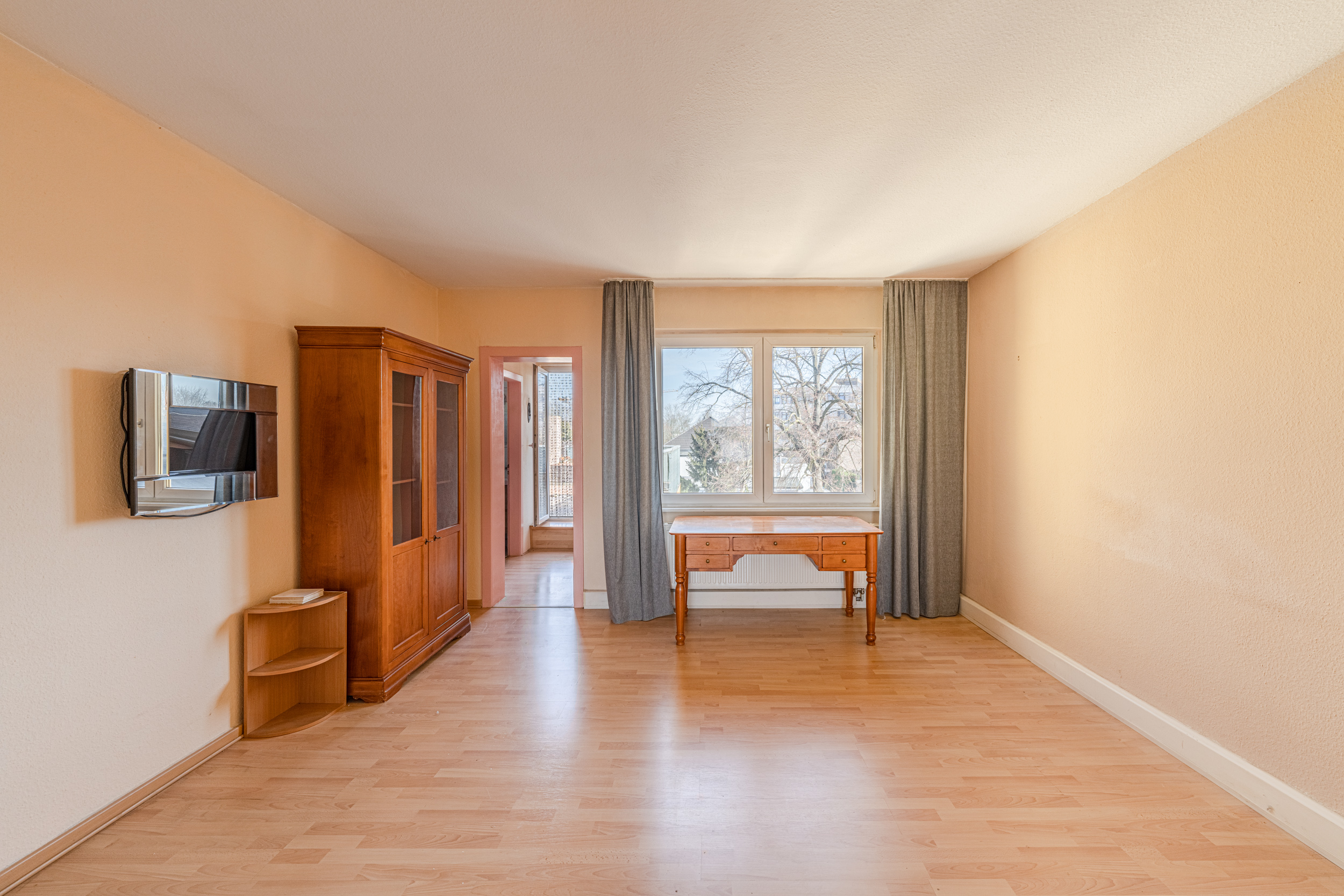Helles Wohnzimmer mit Holzschrank, Schreibtisch am Fenster, Fernseher und Parkettboden.