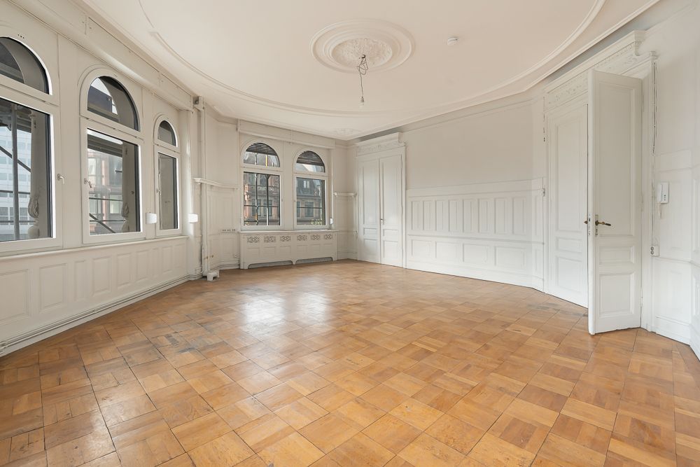 Heller, leerer Raum mit großen Fenstern, weißer Wandvertäfelung und Holzboden.