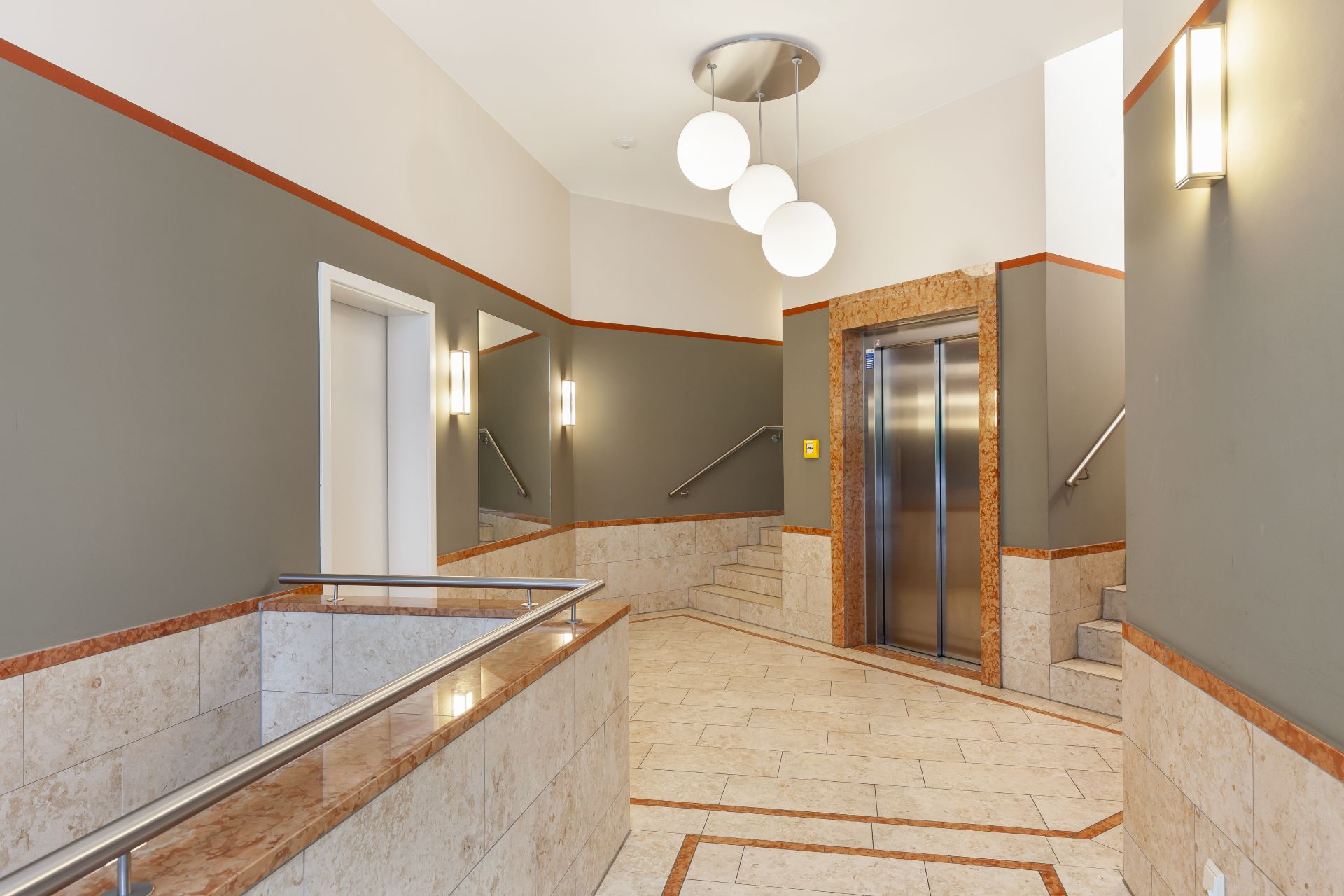 Heller Eingangsbereich mit Aufzug, Rampe, Treppen und Wandlampen in einem Wohngebäude.