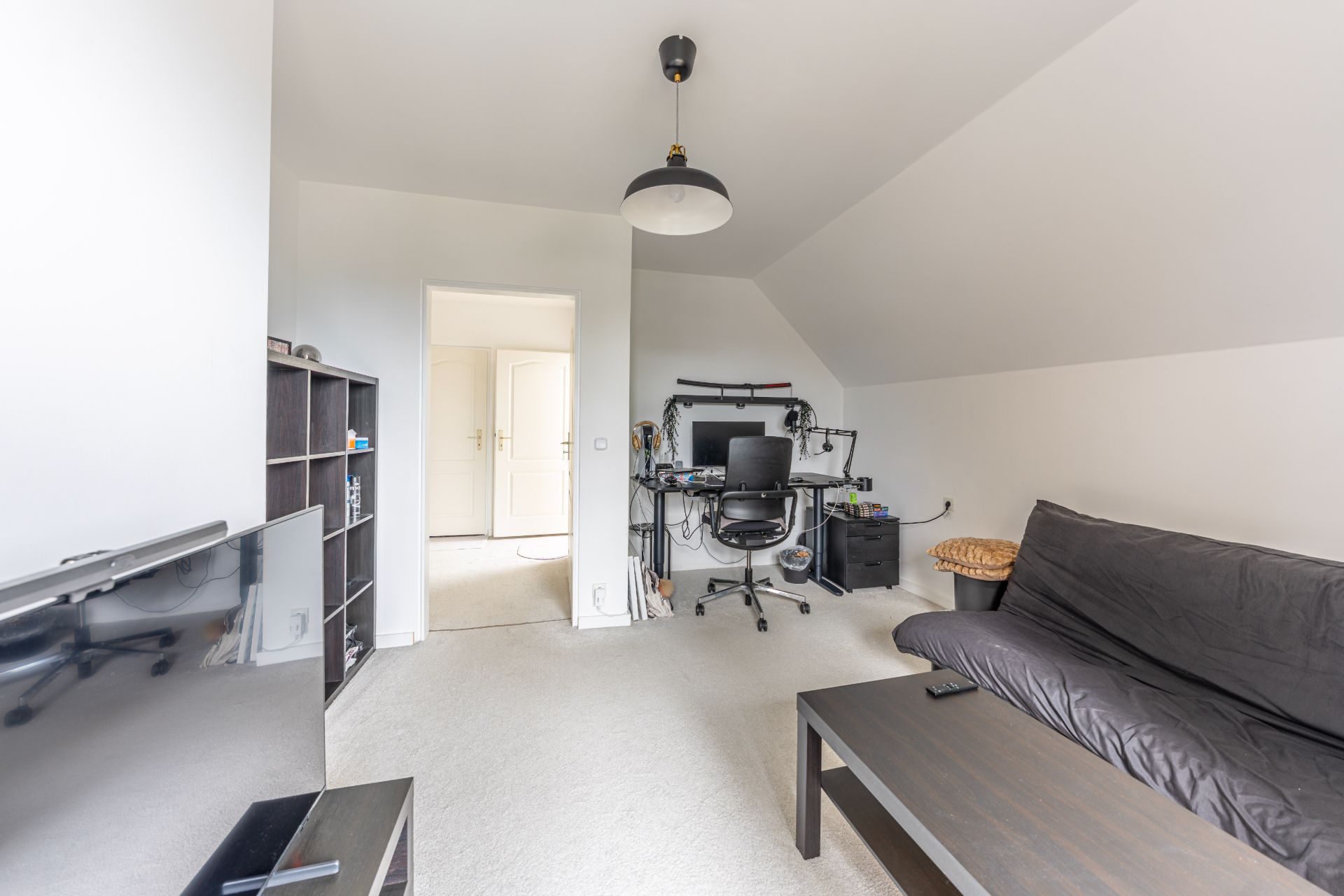 Wohnzimmer mit schwarzem Sofa, Schreibtisch mit Computer, Regal und Fernseher.