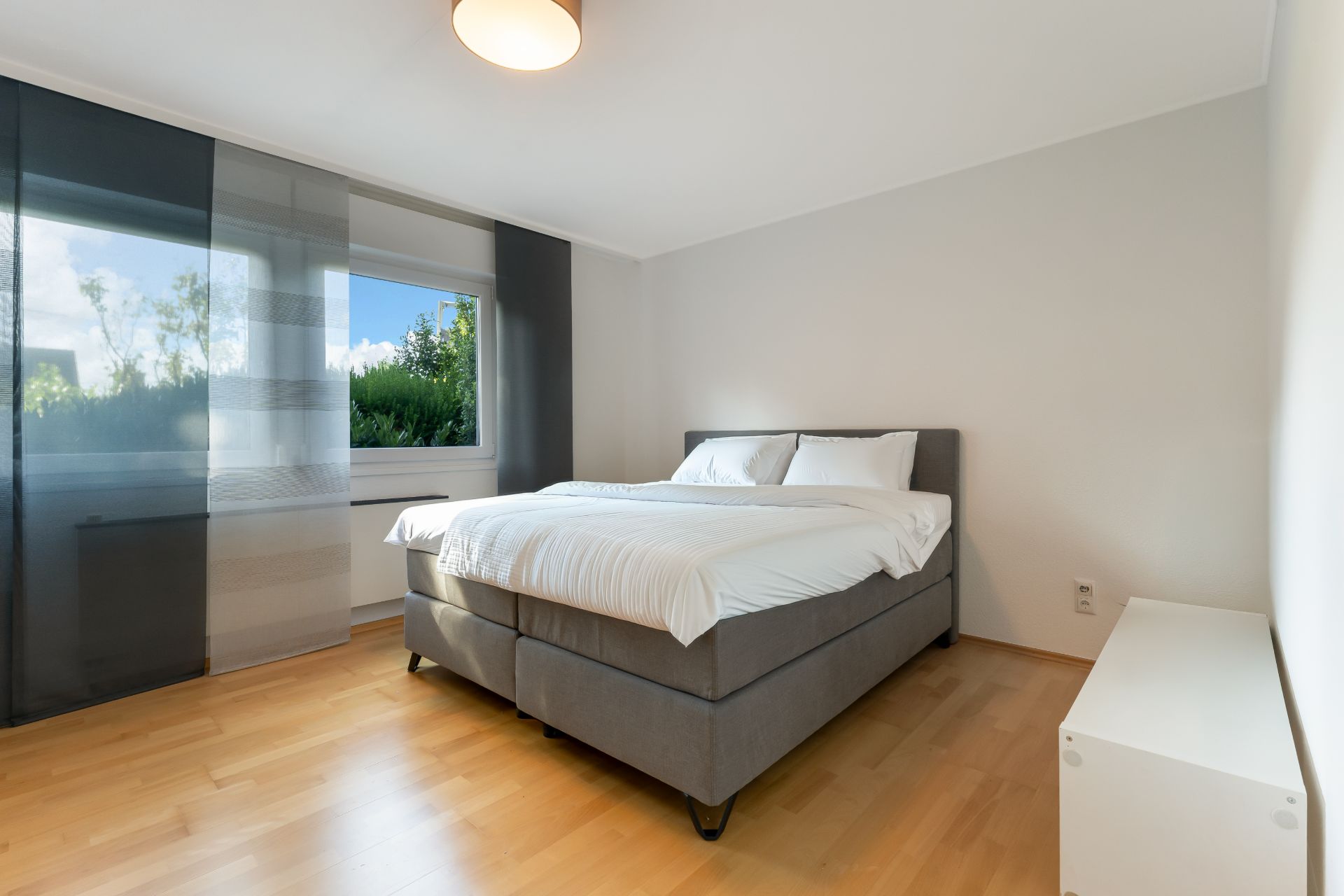 Modernes Schlafzimmer mit Doppelbett, Holzboden und großem Fenster mit Vorhängen.
