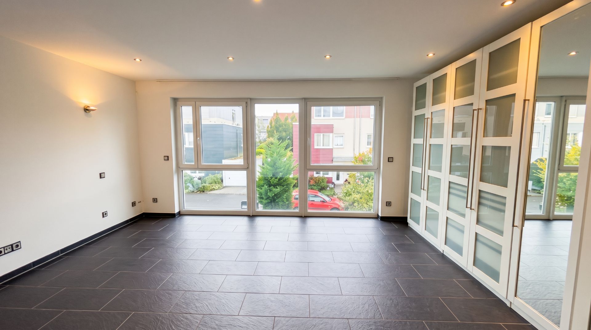 Heller Raum mit großen Fenstern, dunklem Fliesenboden und einem Einbauschrank mit Glastüren.