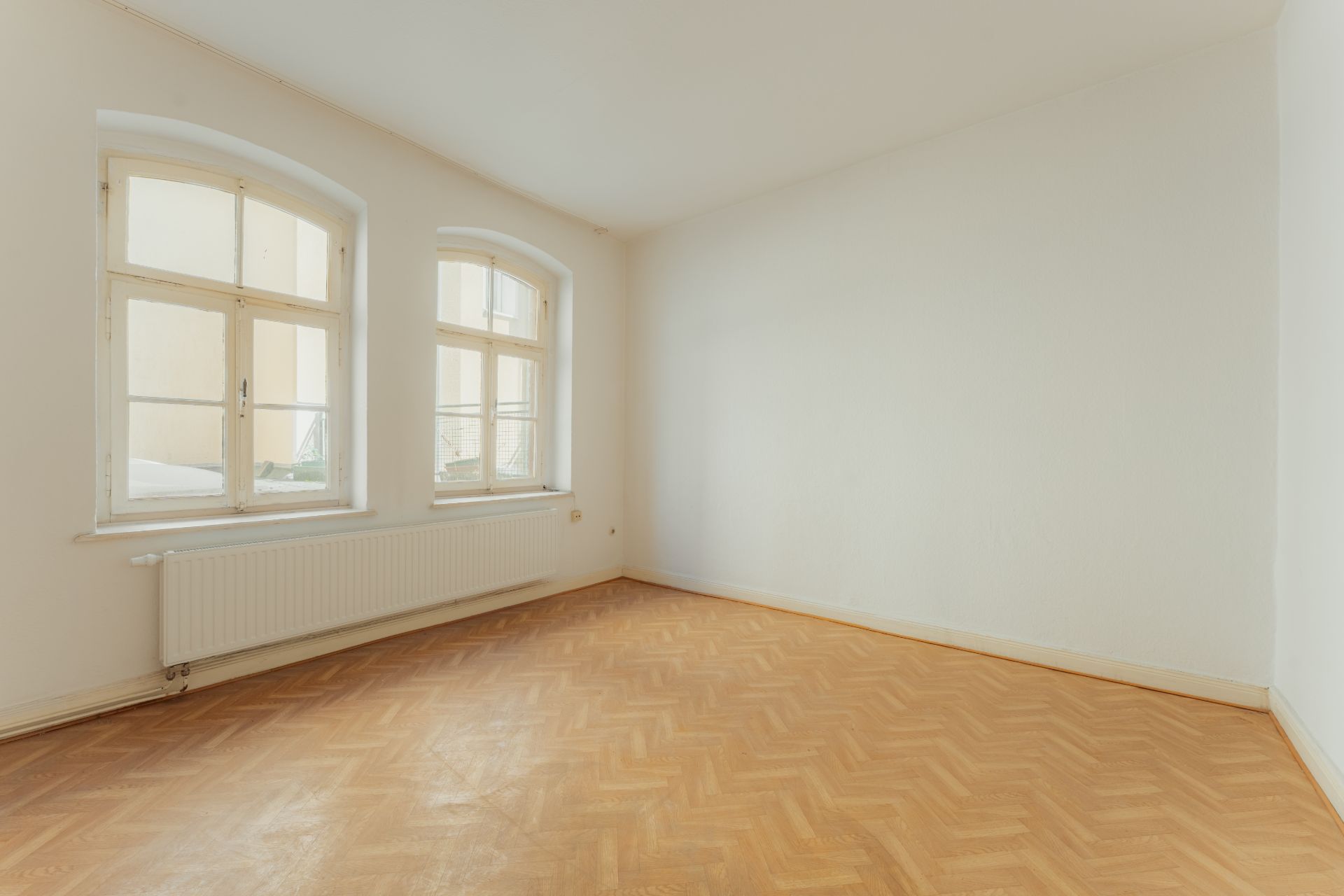 Leerer Raum mit Parkettboden, weißen Wänden und zwei großen Fenstern.