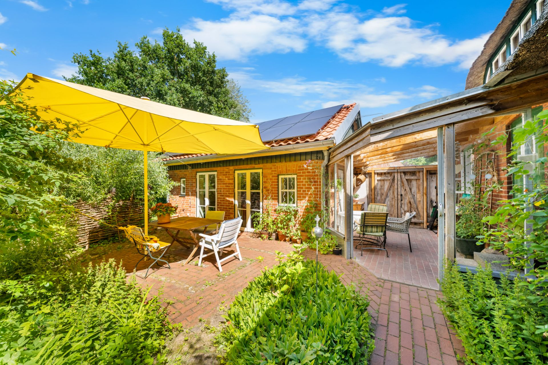 Gemütliche Terrasse mit gelbem Sonnenschirm, Sitzmöbeln und Ziegelhaus im Grünen.