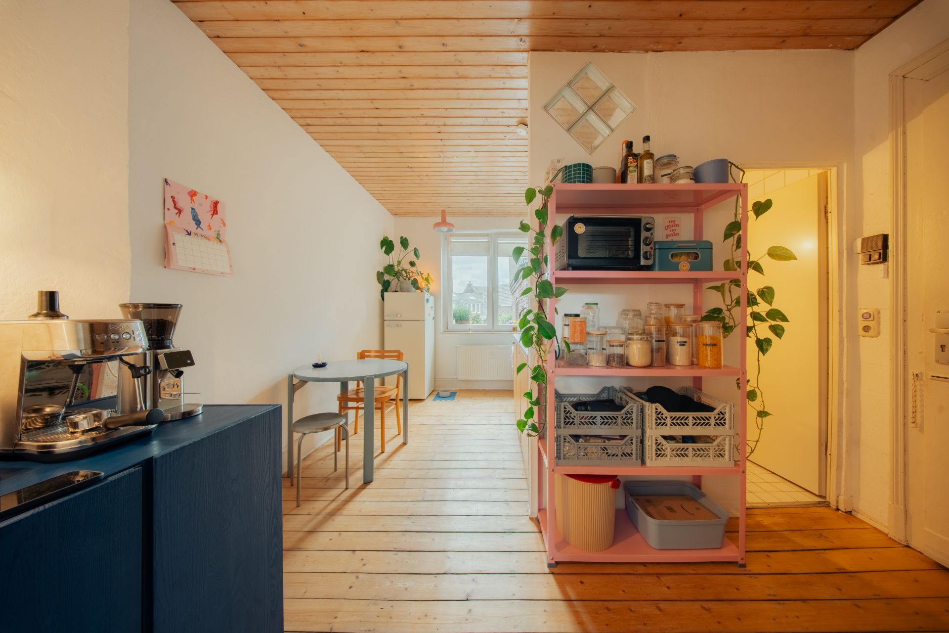 Helle Küche mit Holzdecke, offenem Regal, Tisch, Stühlen, Pflanze und Kaffeemaschine.