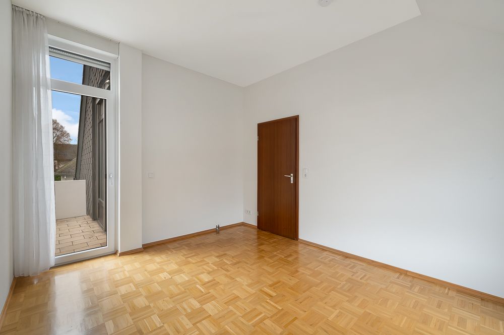 Leeres Zimmer mit Parkettboden, weißer Wand, Holztür und Zugang zu einem Balkon.