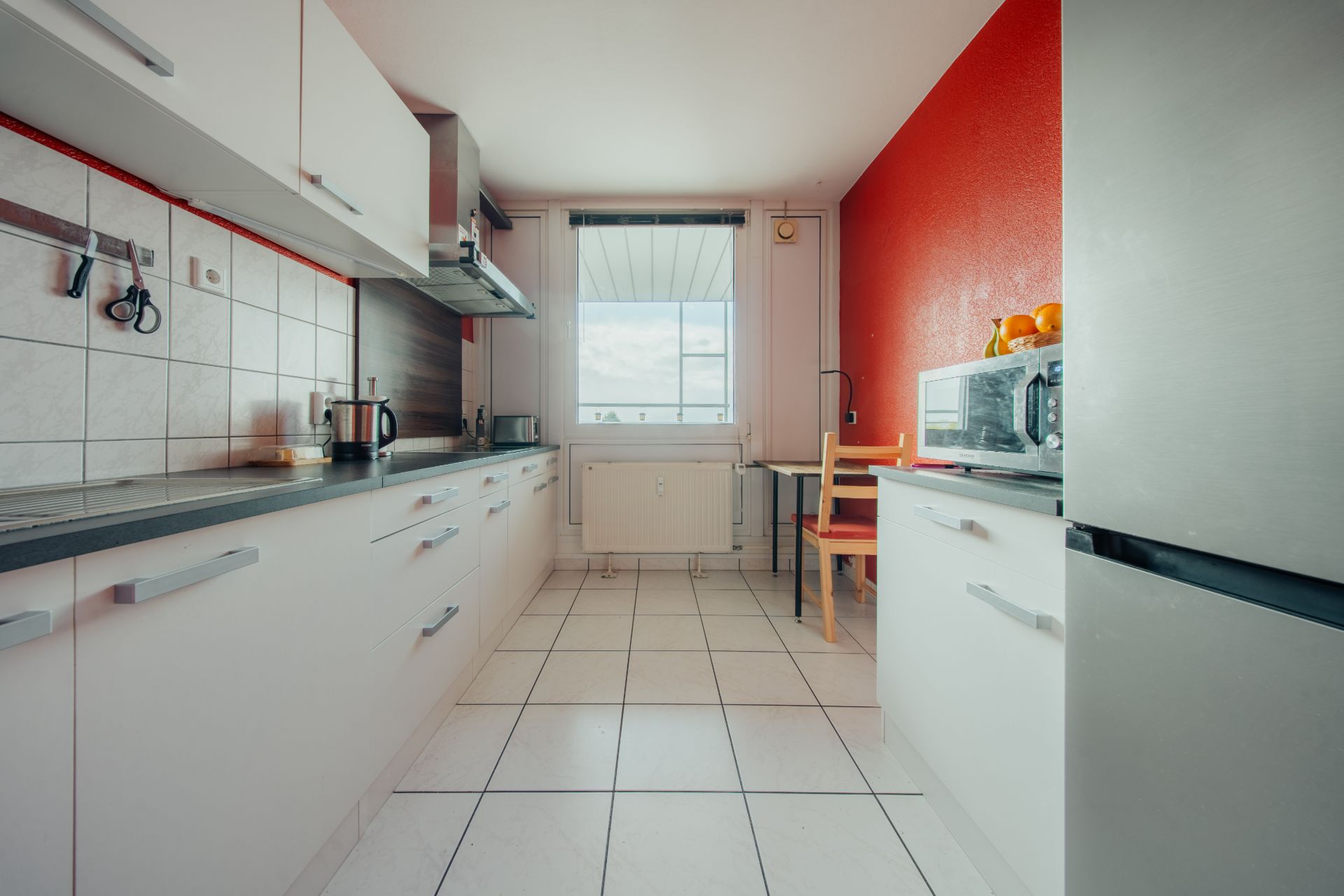 Helle Küche mit weißen Schränken, roter Wand, großem Fenster und Esstisch mit zwei Stühlen.