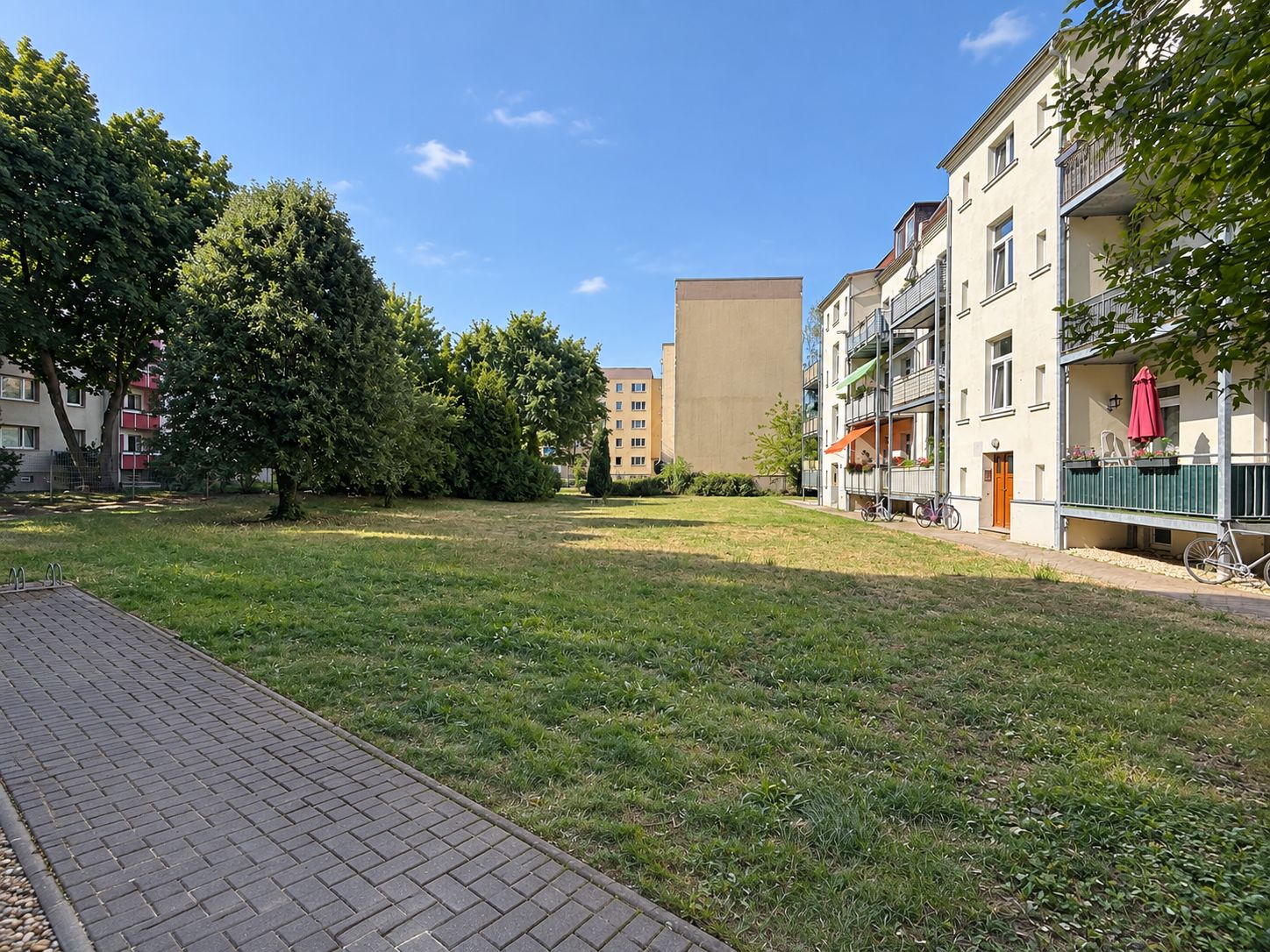 Grüne Wiese mit Gehweg, umgeben von Wohnhäusern und Bäumen an einem sonnigen Tag.