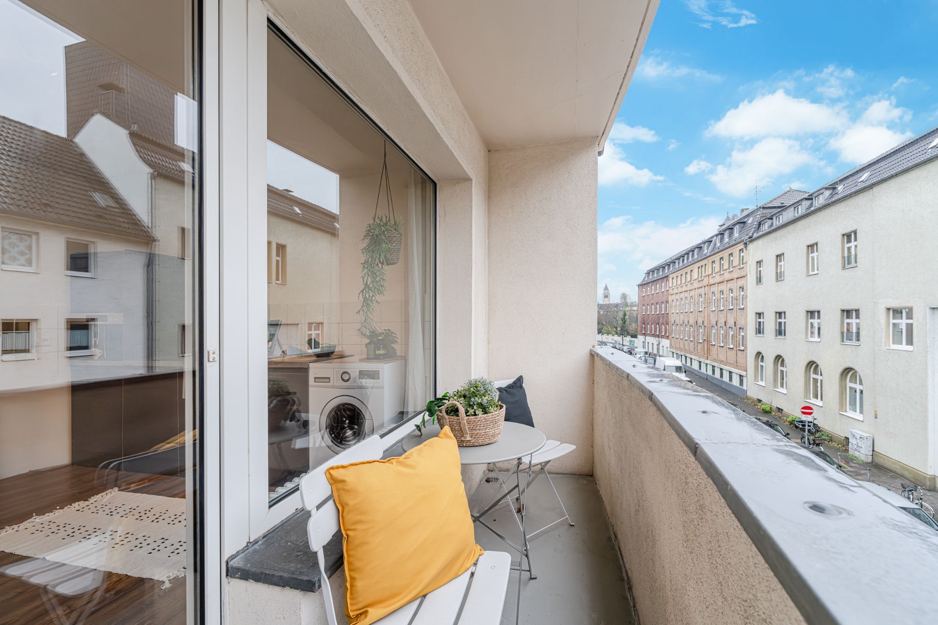 Balkon mit weißer Bank, gelbem Kissen, kleinem Tisch und Blick auf eine ruhige Wohnstraße.