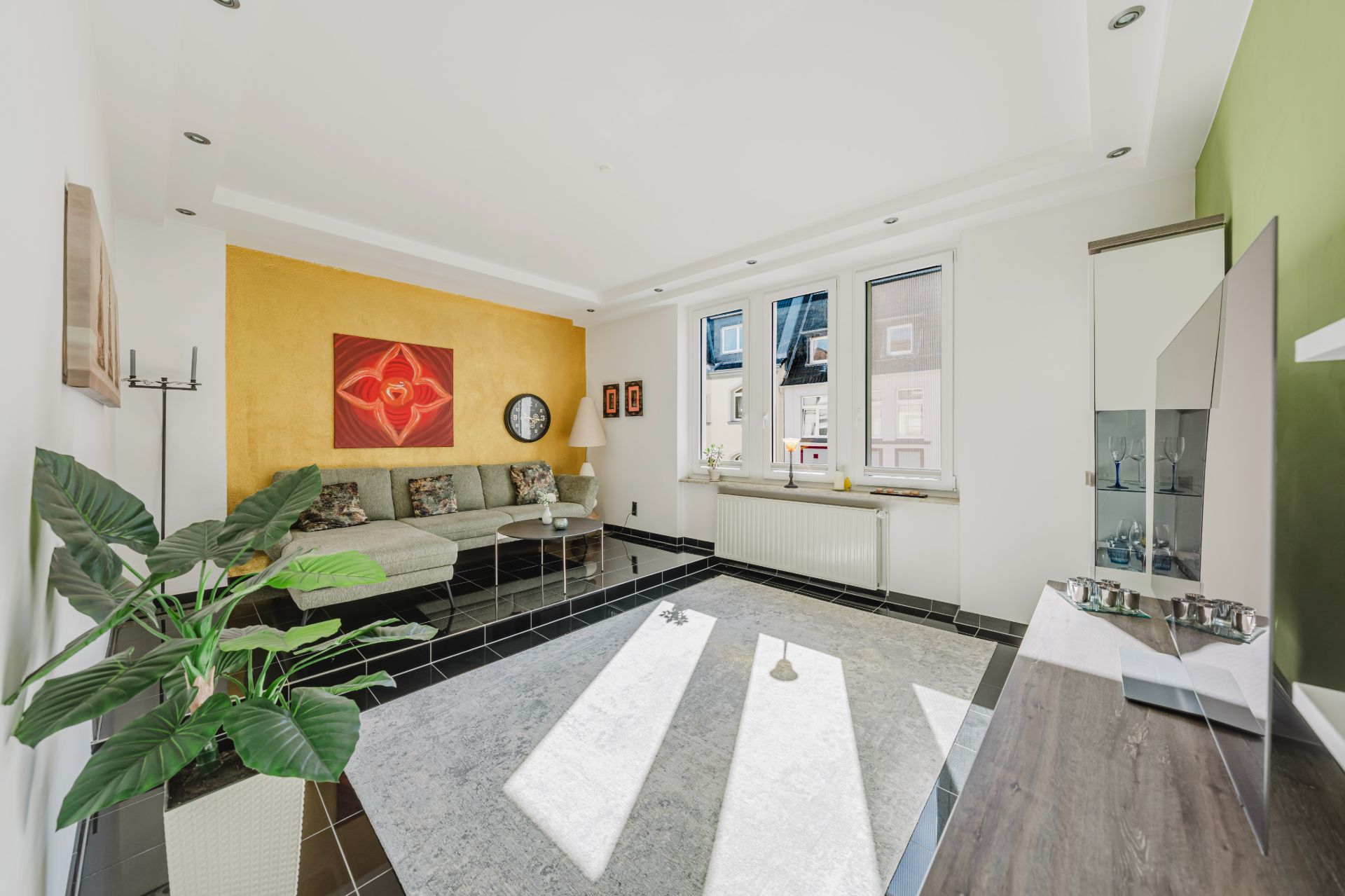 Helles Wohnzimmer mit Ecksofa, großem Teppich, Pflanze und gelber Wand mit Kunstbild.