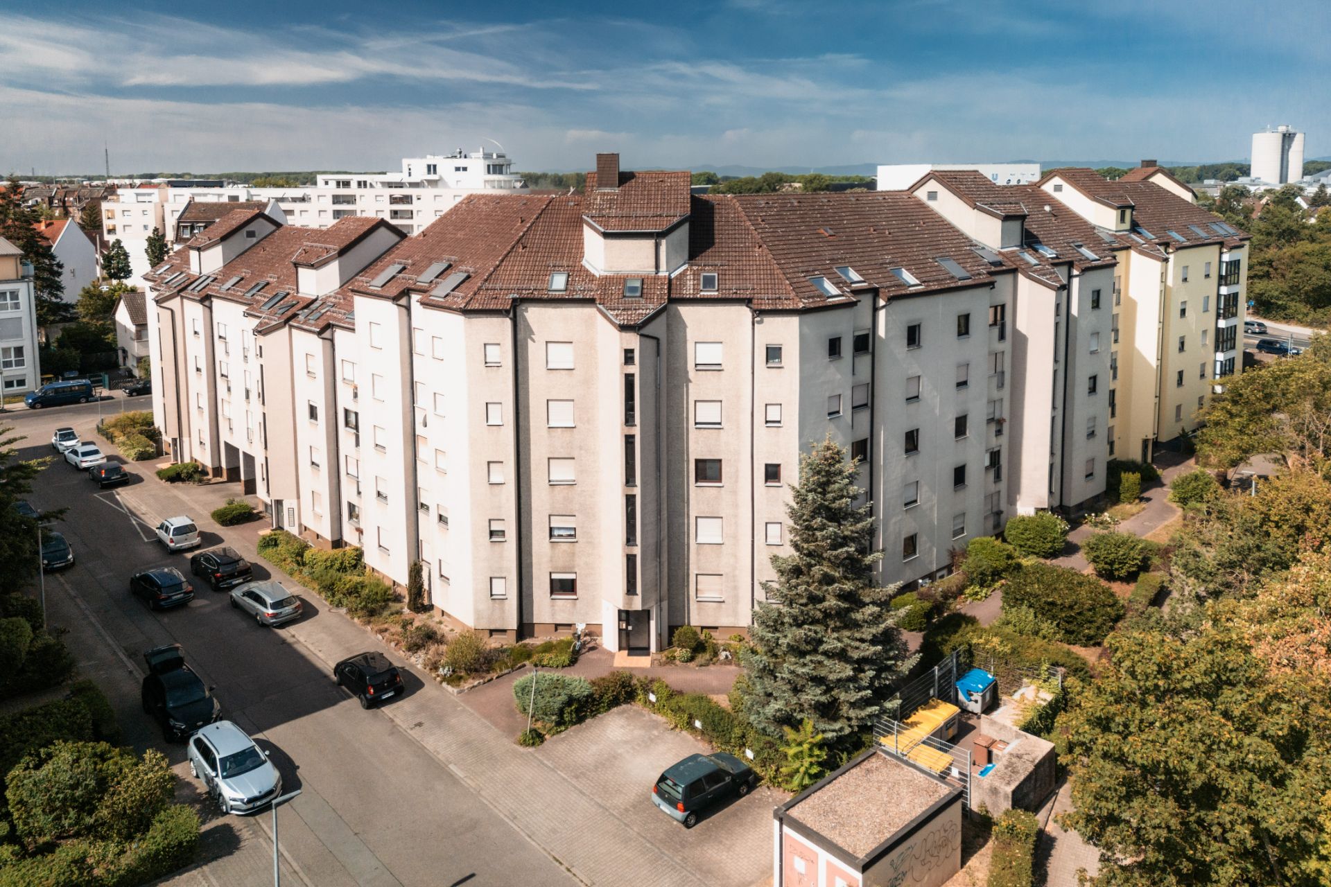 Mehrstöckiges Wohnhaus mit braunem Dach, umgeben von Autos und Bäumen an einer ruhigen Straße.