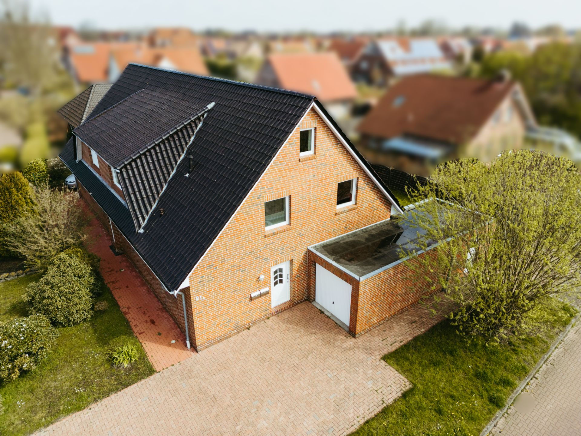 Backsteinhaus mit schwarzem Dach, Garage und gepflasterter Einfahrt, umgeben von Büschen.