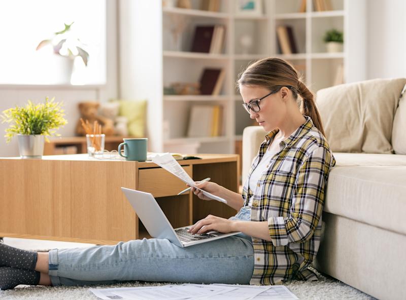 Frau sitzt auf Teppich, arbeitet am Laptop und liest Papiere in modernem Wohnzimmer.