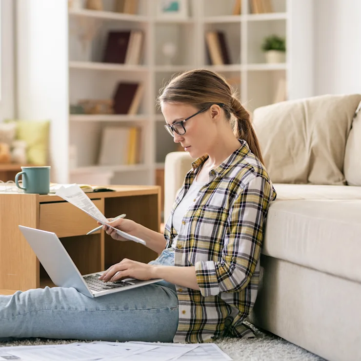 Frau sitzt auf Teppich, arbeitet am Laptop und liest Papiere in modernem Wohnzimmer.