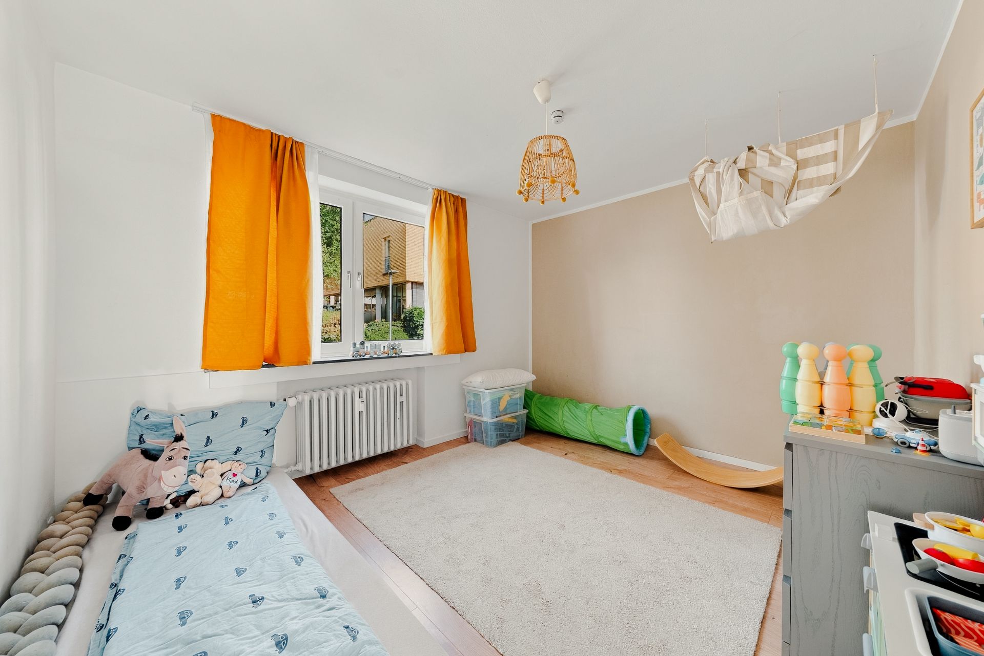 Helles Kinderzimmer mit Einzelbett, bunten Vorhängen und Spielzeug auf einem Teppich.