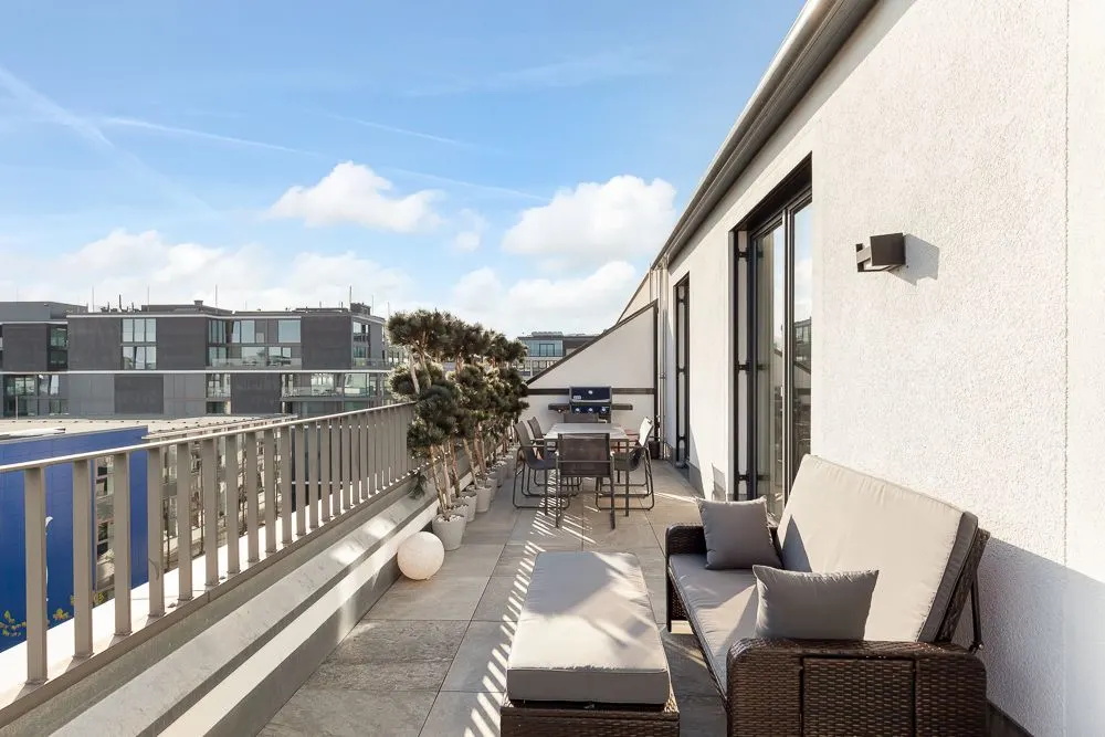 Große Dachterrasse mit Sitzbank, Tisch, Stühlen und Pflanzen, umgeben von modernen Gebäuden.