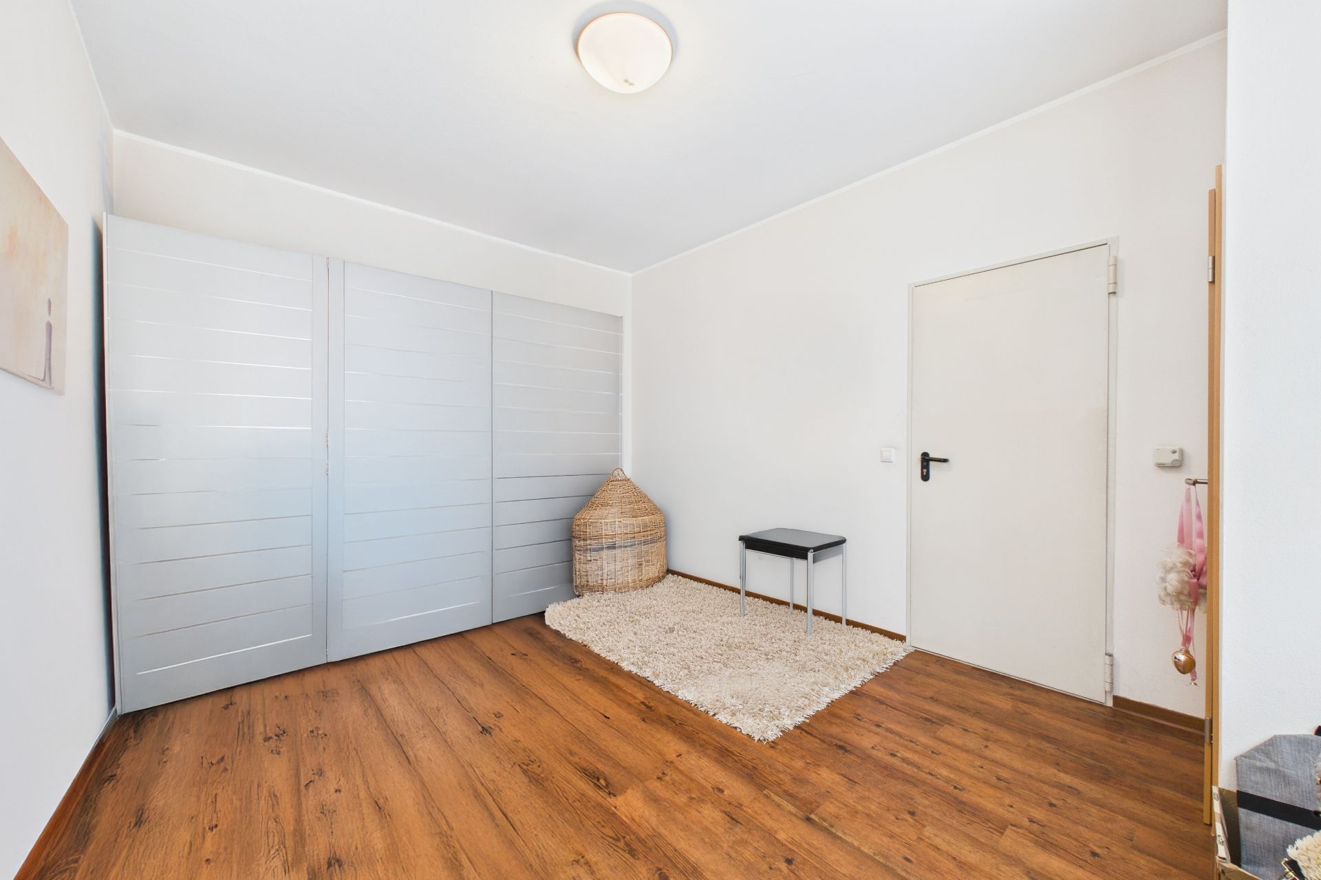 Helles Zimmer mit Holzfußboden, Teppich, großem Schrank und kleinem Hocker neben einer Tür.