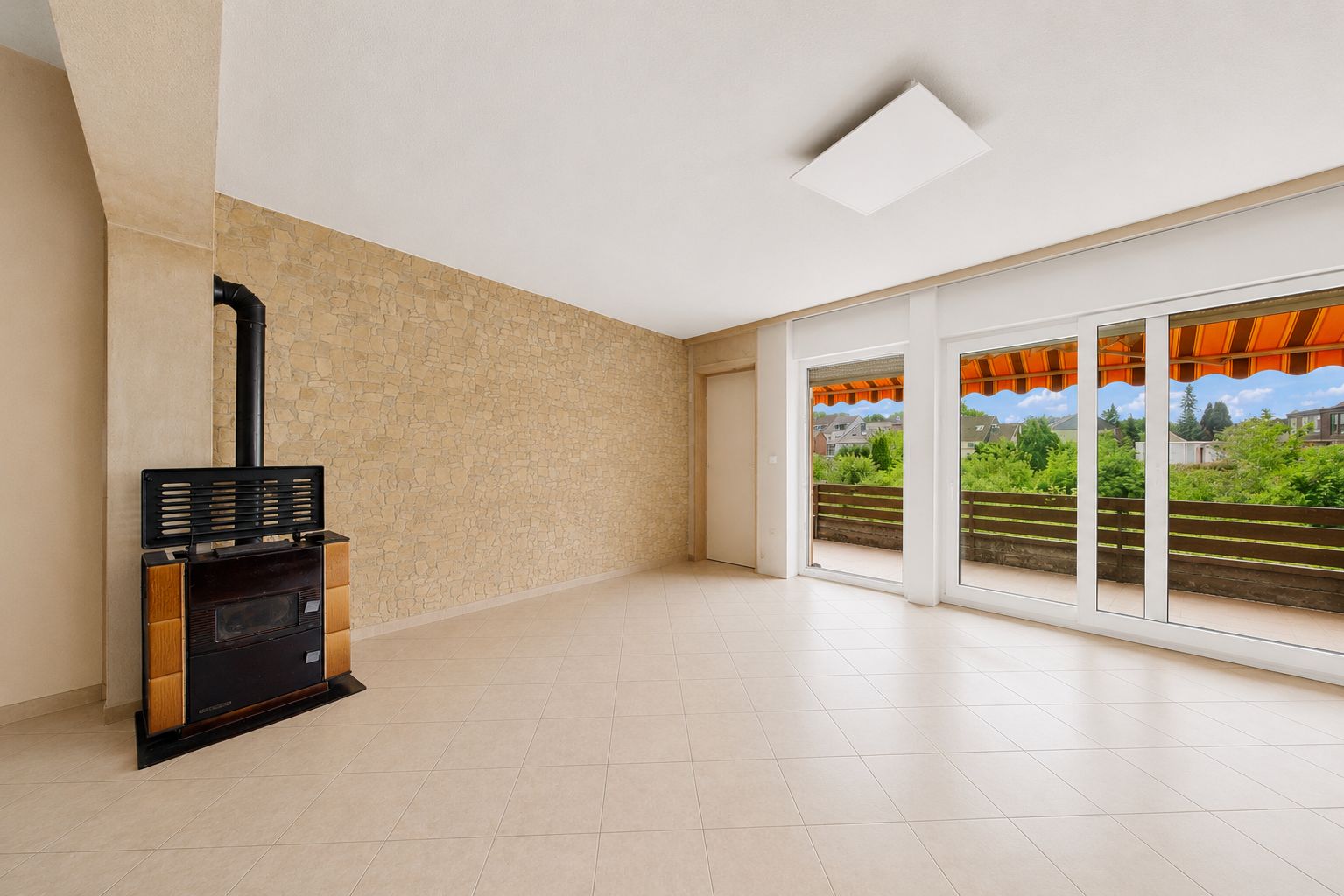 Leeres, helles Wohnzimmer mit Fliesenboden, Kamin und großen Fenstern zur Terrasse.