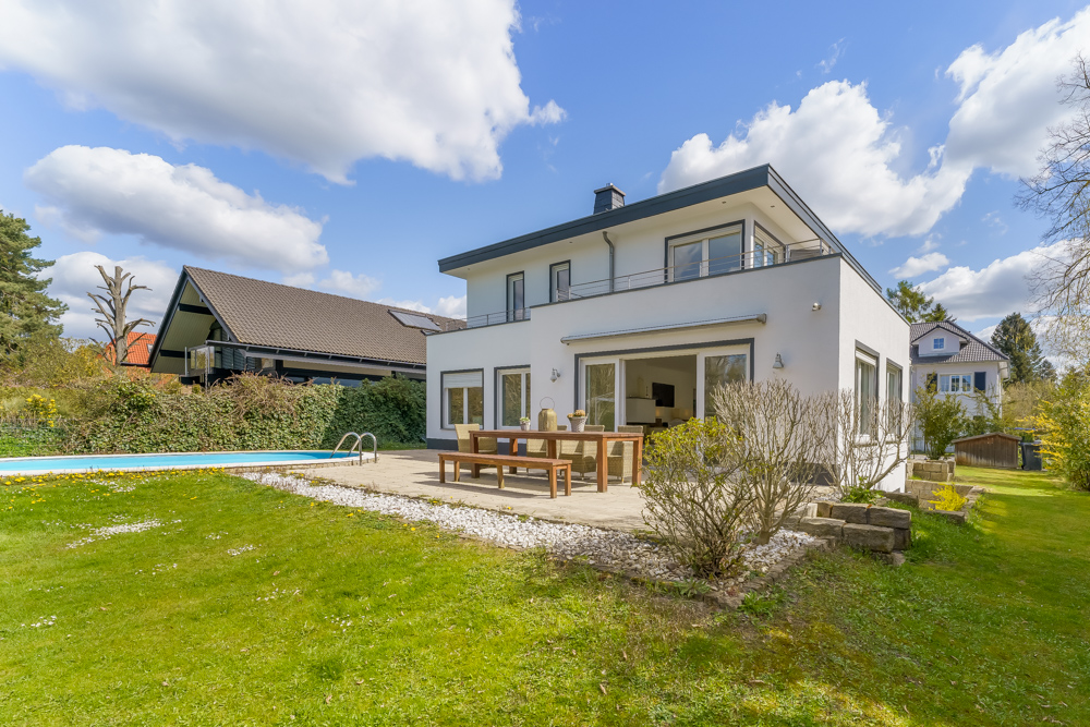 Modernes weißes Wohnhaus mit Terrasse, Holztisch, Garten und Pool an einem sonnigen Tag.