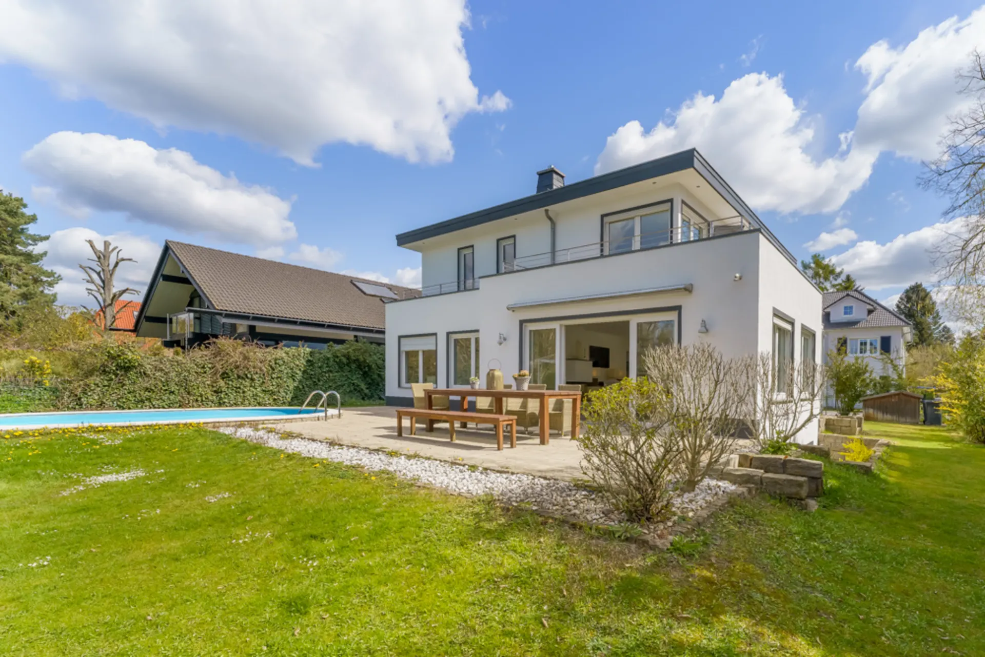 Modernes weißes Wohnhaus mit Terrasse, Holztisch, Garten und Pool an einem sonnigen Tag.