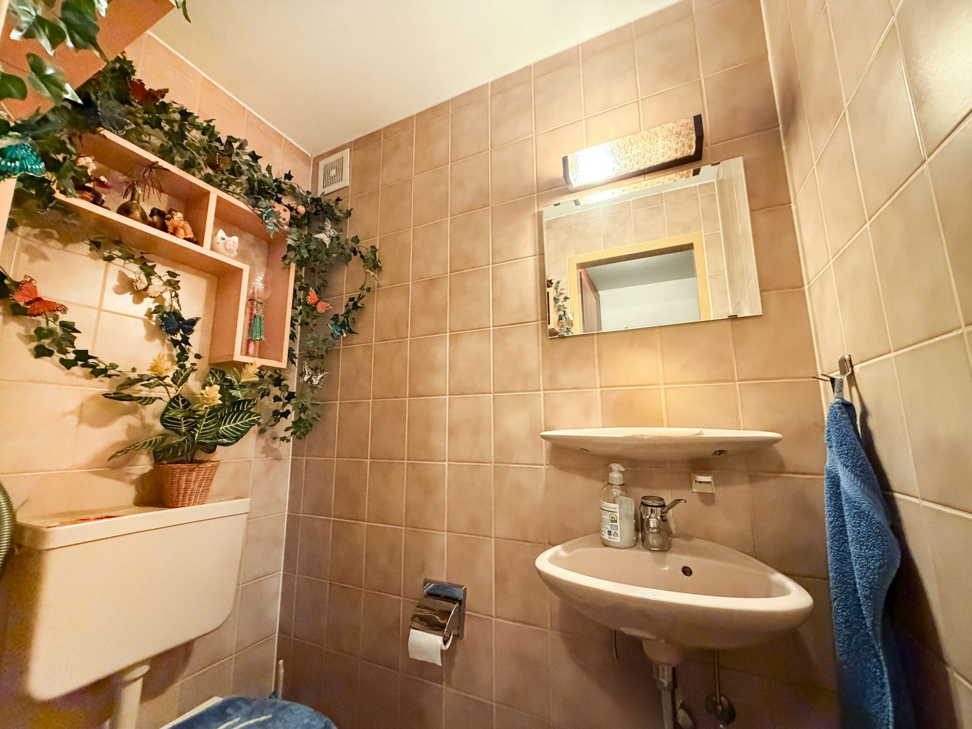 Kleines Badezimmer mit beigem Fliesenspiegel, Waschbecken, Spiegel und Dekoration mit Kunstpflanzen.