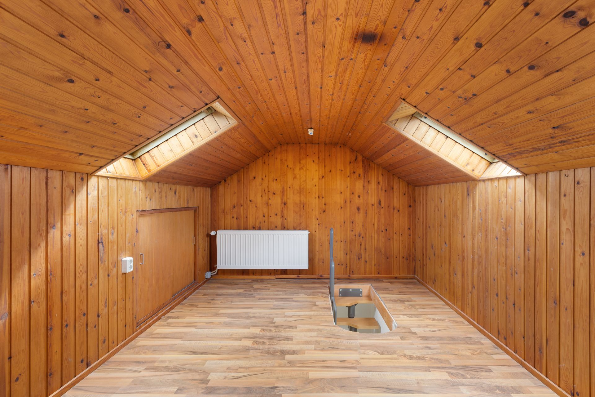 Dachzimmer mit Holzverkleidung, zwei Dachfenstern, Heizkörper und Treppenöffnung im Boden.