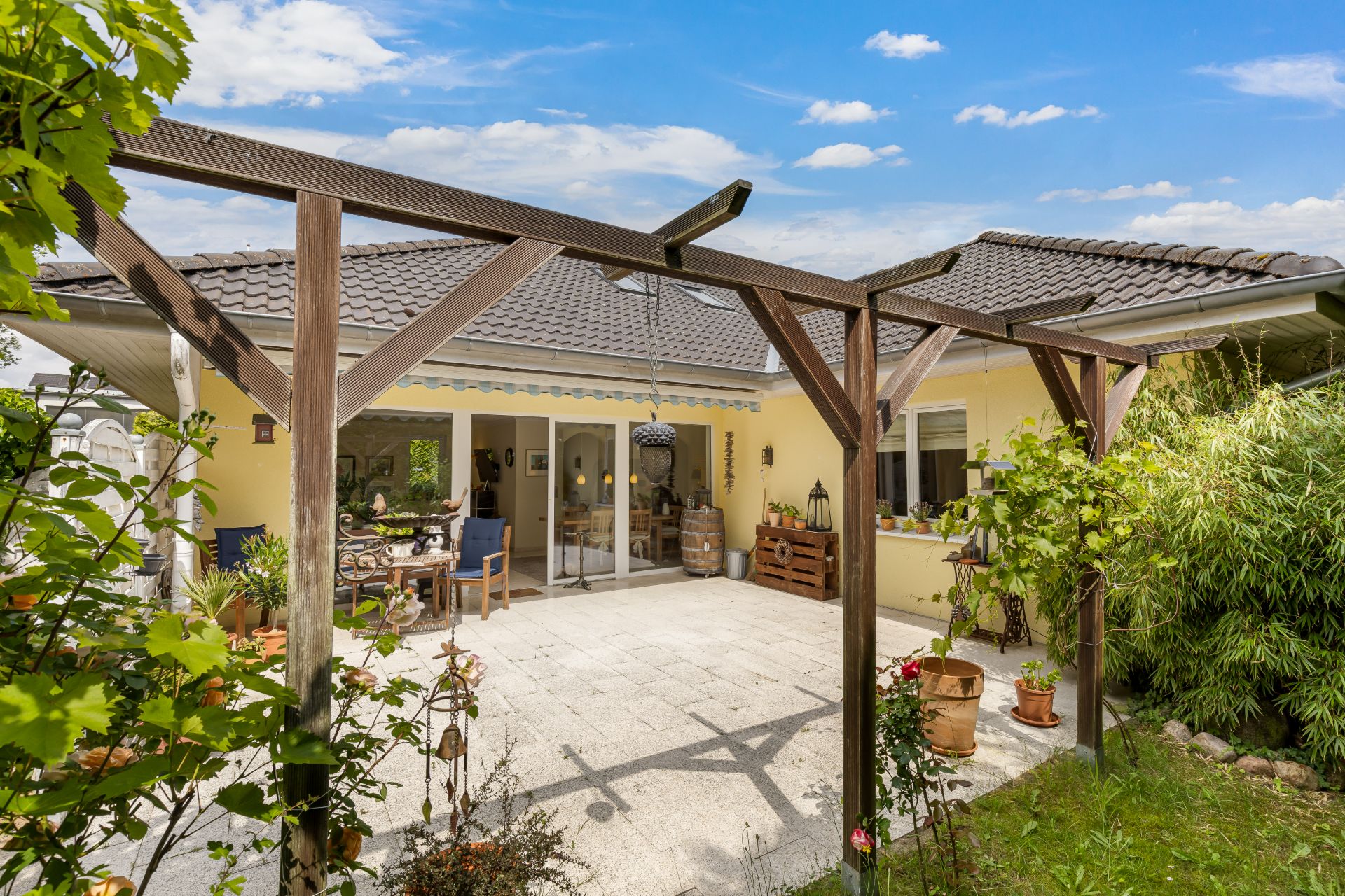 Terrasse mit Holzbalken, Pflanzkübeln und Sitzbereich vor einem gelben Haus.