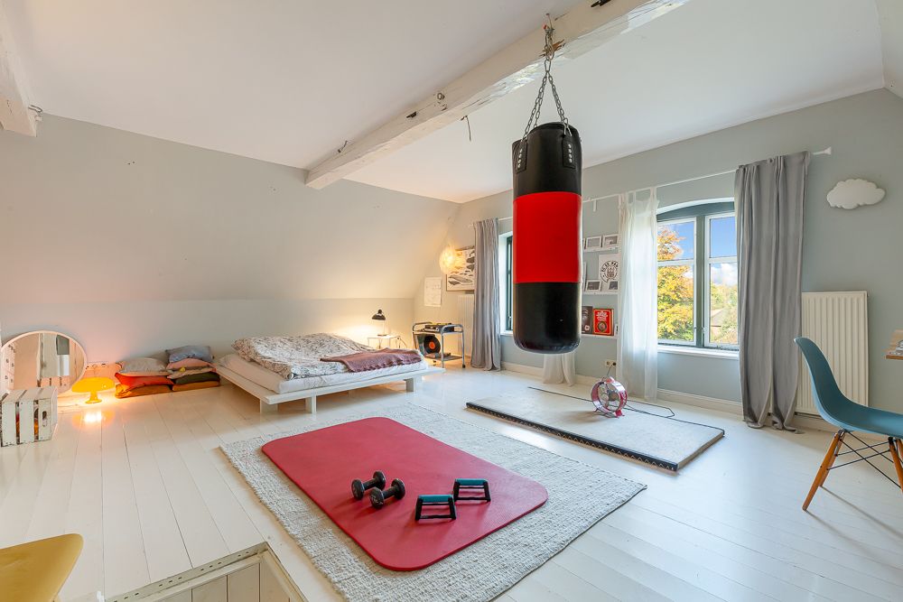 Helles Schlafzimmer mit Bett, Boxsack, Fenster, Fitnessmatte und Hanteln auf weißem Holzboden.
