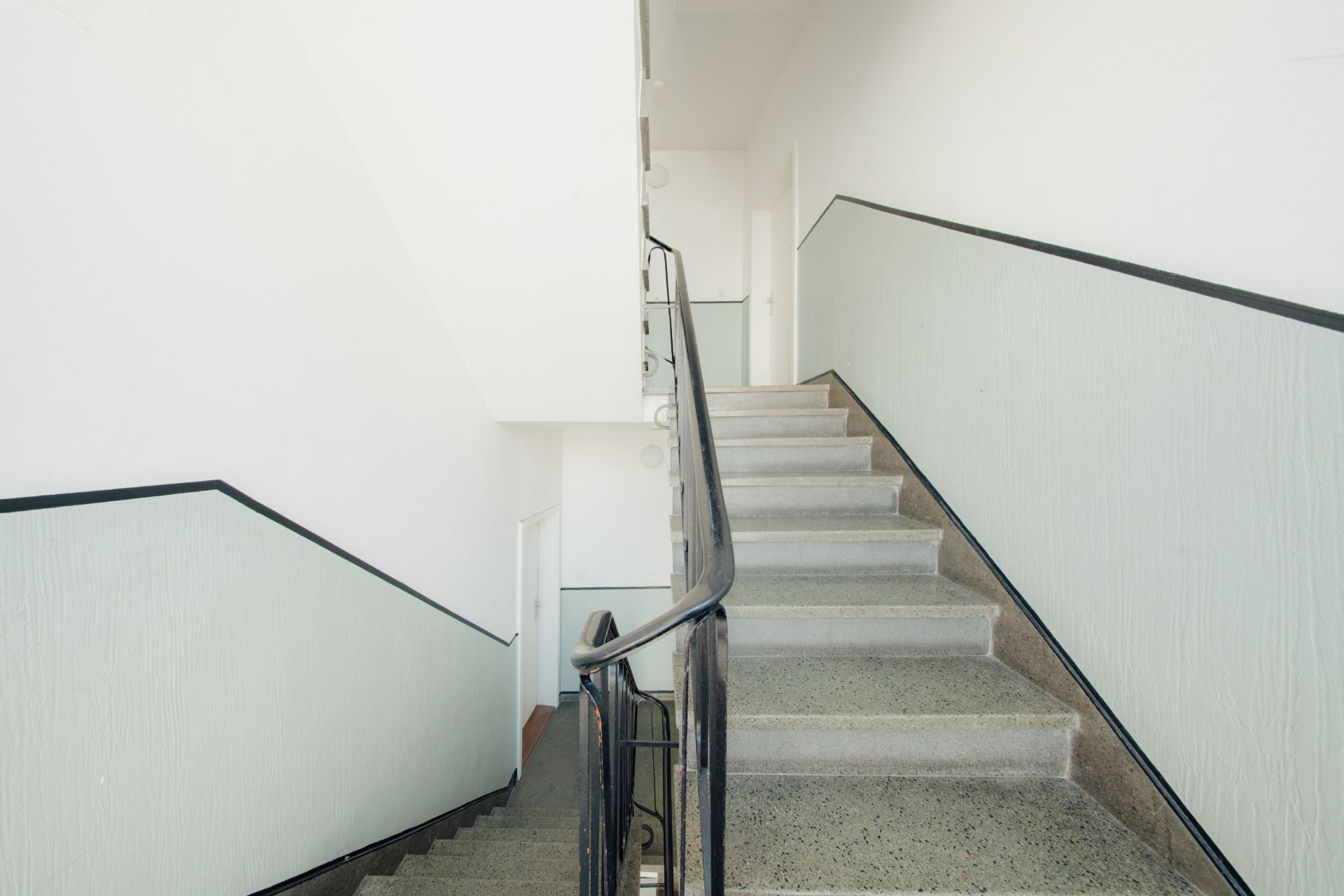 Helles Treppenhaus mit Metallgeländer und hellgrünen Wänden in einem Wohngebäude.