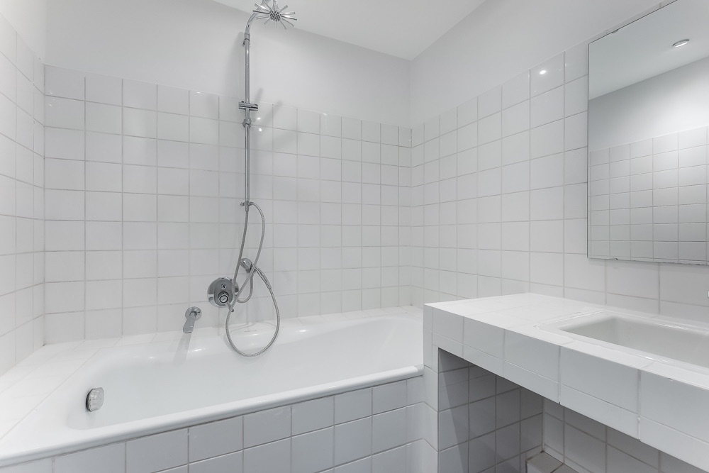 Modernes weiß gefliestes Bad mit Badewanne, Duschkopf, Waschbecken und Spiegel.