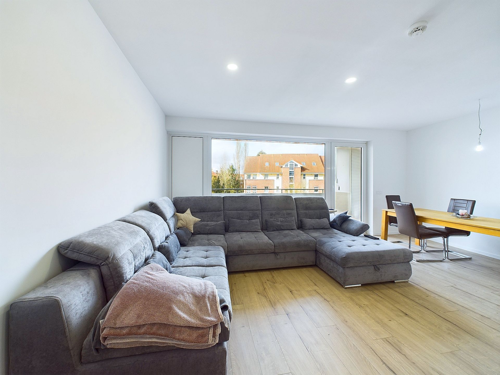 Helles Wohnzimmer mit großem grauen Ecksofa, Holzboden und Esstisch vor großem Fenster.