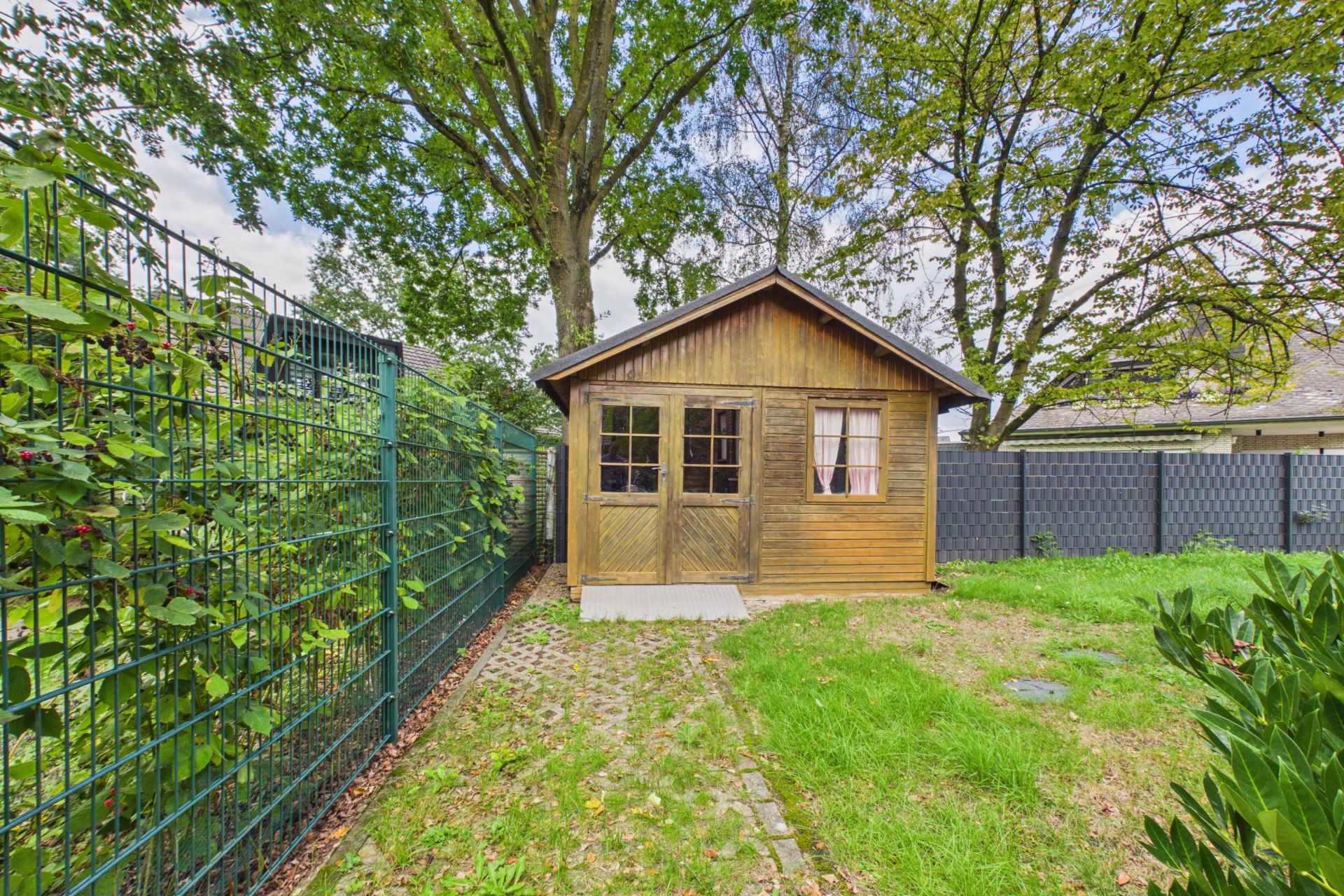 Kleines Holzhaus im Garten, umgeben von Rasen und Bäumen, mit Zaun links.