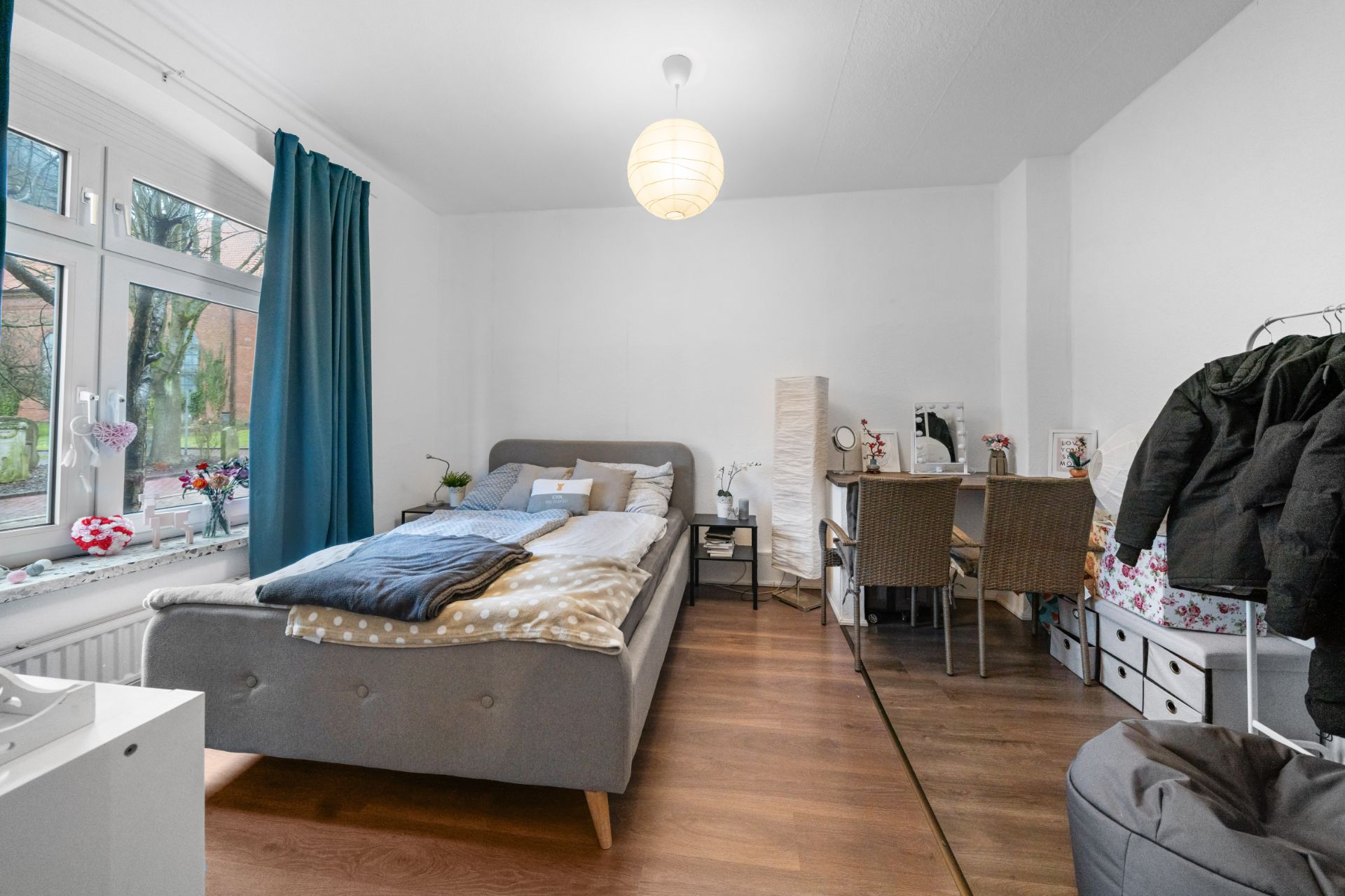 Helles Schlafzimmer mit Doppelbett, Schreibtisch, Fenster und Garderobe auf Holzfußboden.
