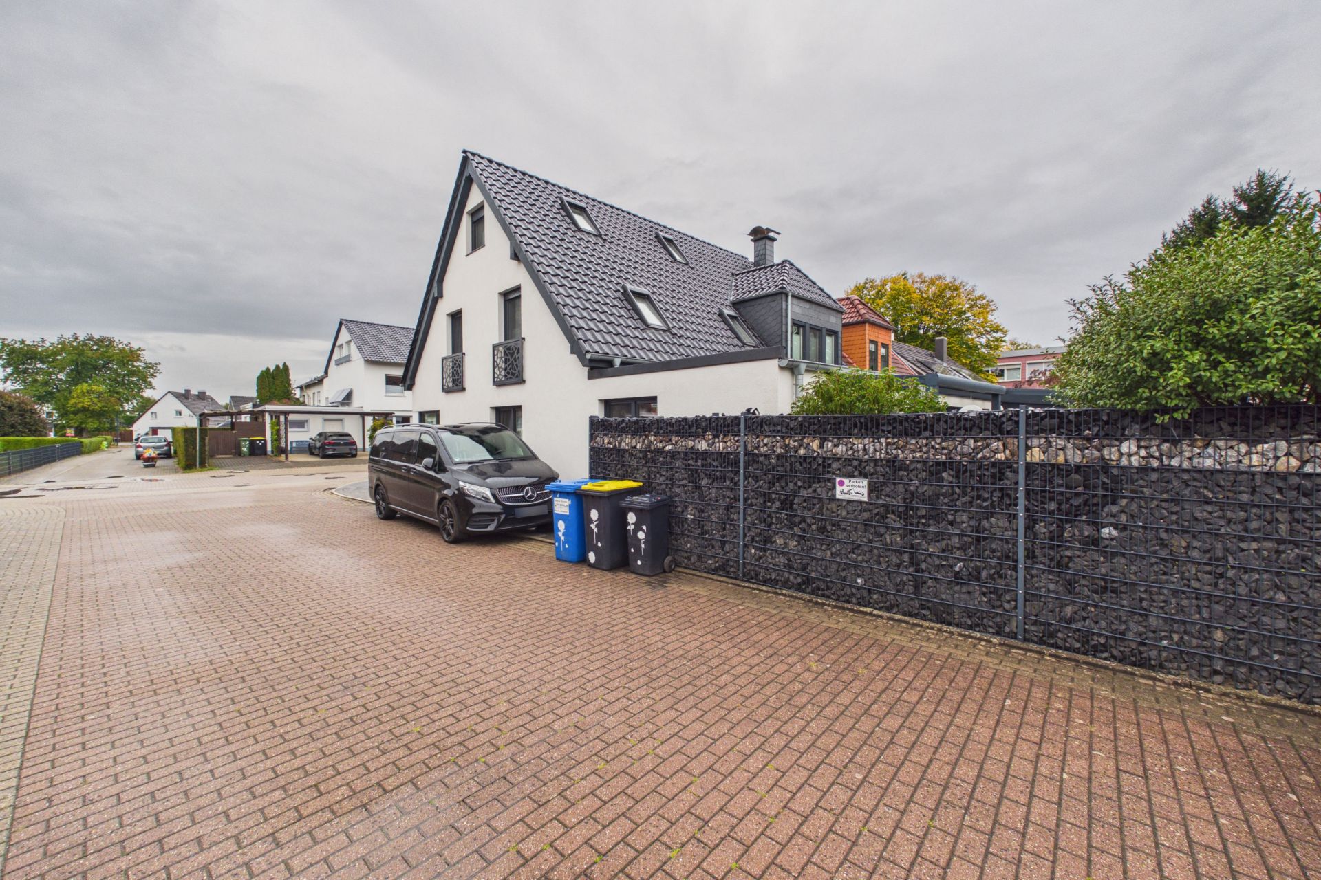 Modernes weißes Wohnhaus mit schwarzem Dach, schwarzem Auto und Mülltonnen auf gepflastertem Hof.