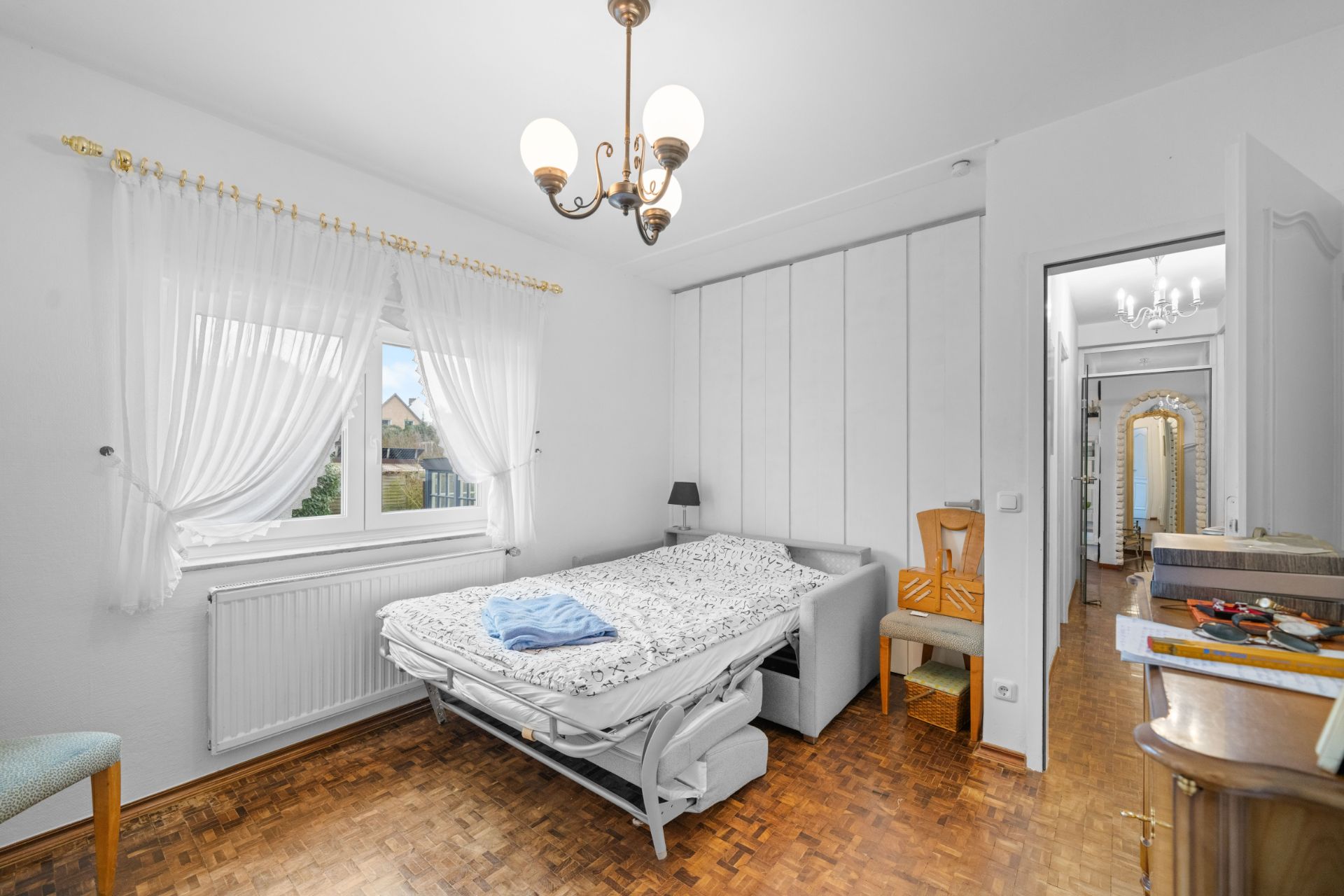 Helles Schlafzimmer mit großem Fenster, Doppelbett, Stuhl und kleinem Nachttisch.