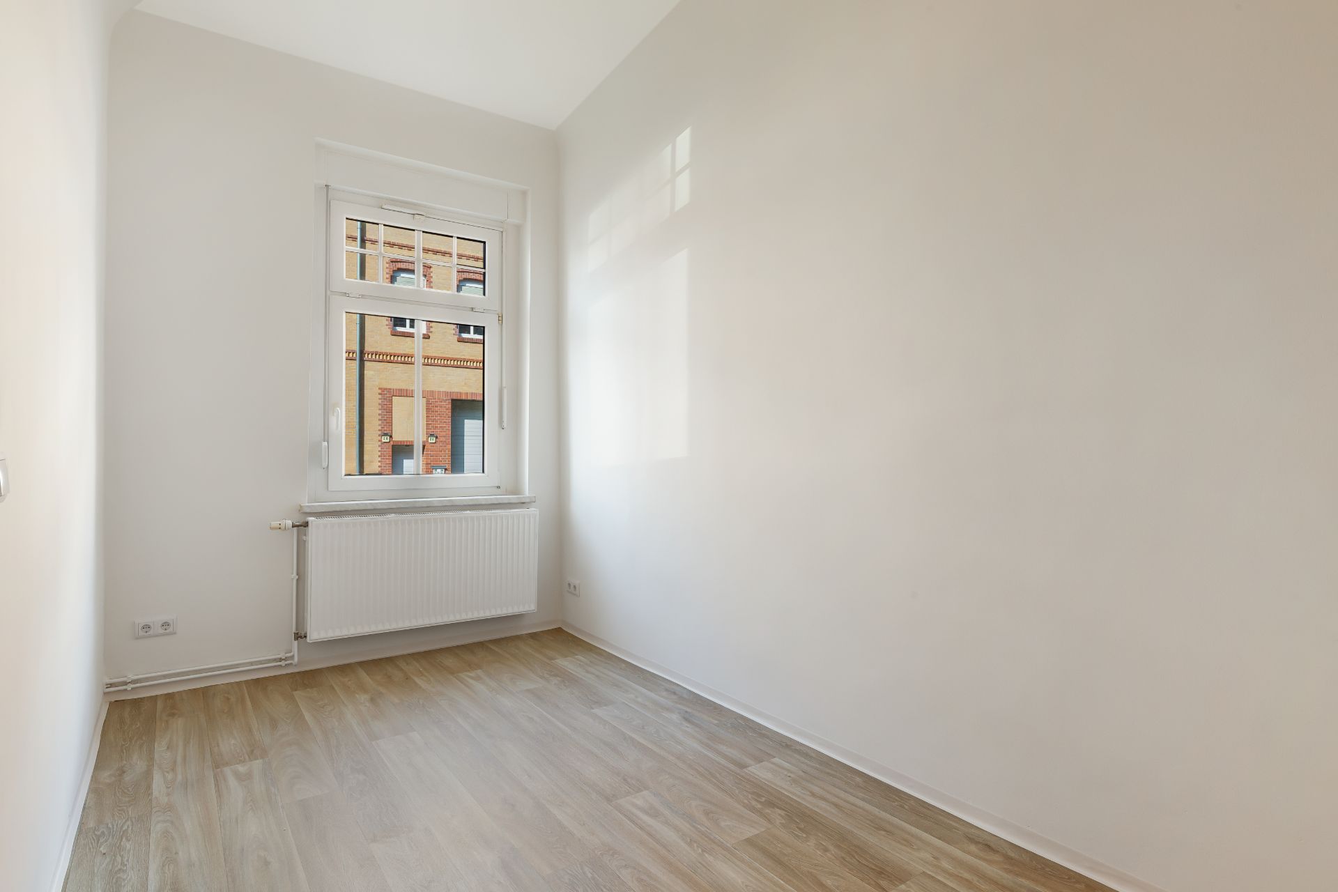 Kleiner leerer Raum mit hellem Holzboden, weißer Wand und Fenster mit Heizkörper.