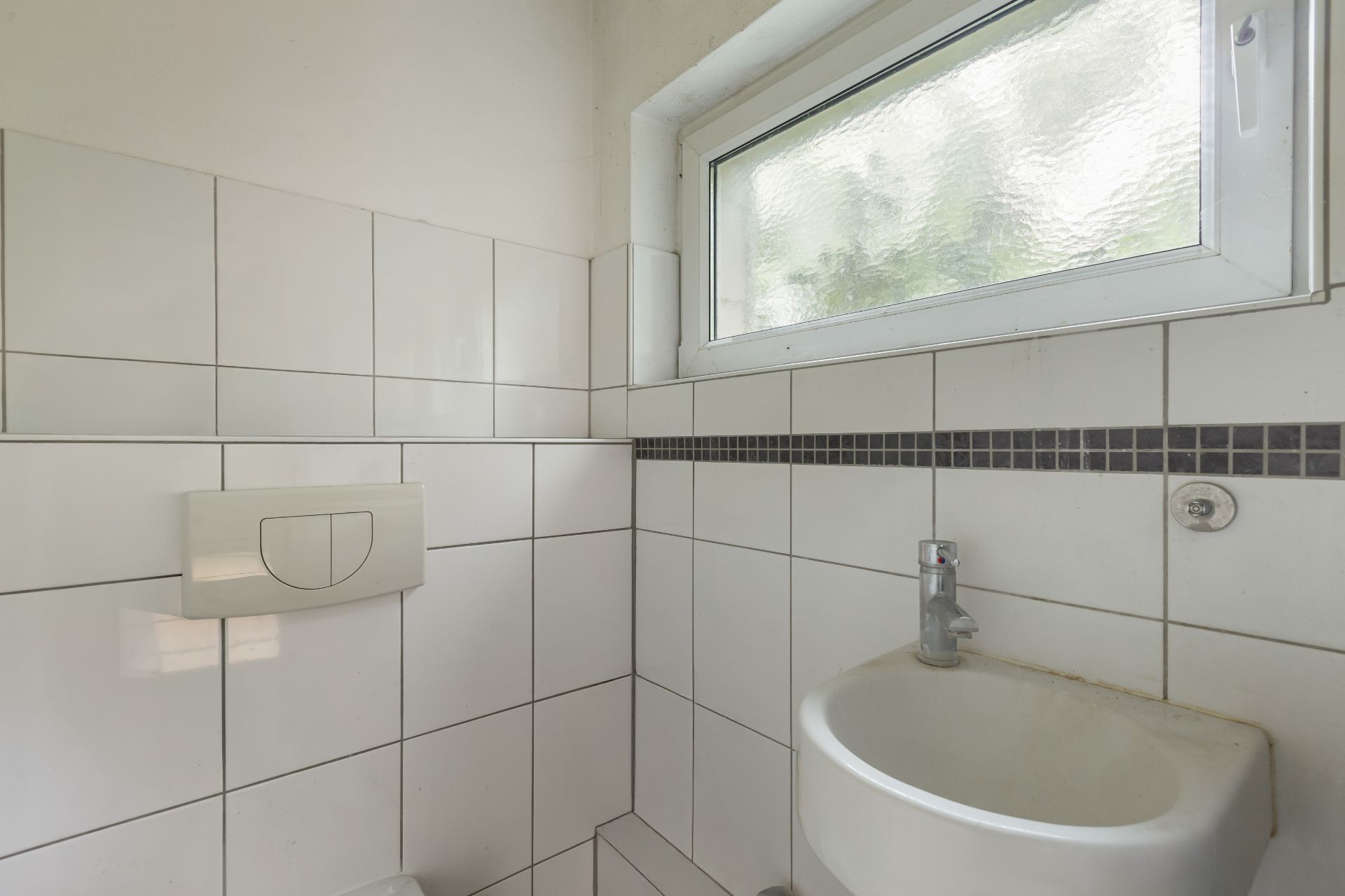 Kleines Badezimmer mit weiß gefliester Wand, kleinem Waschbecken und Fenster.