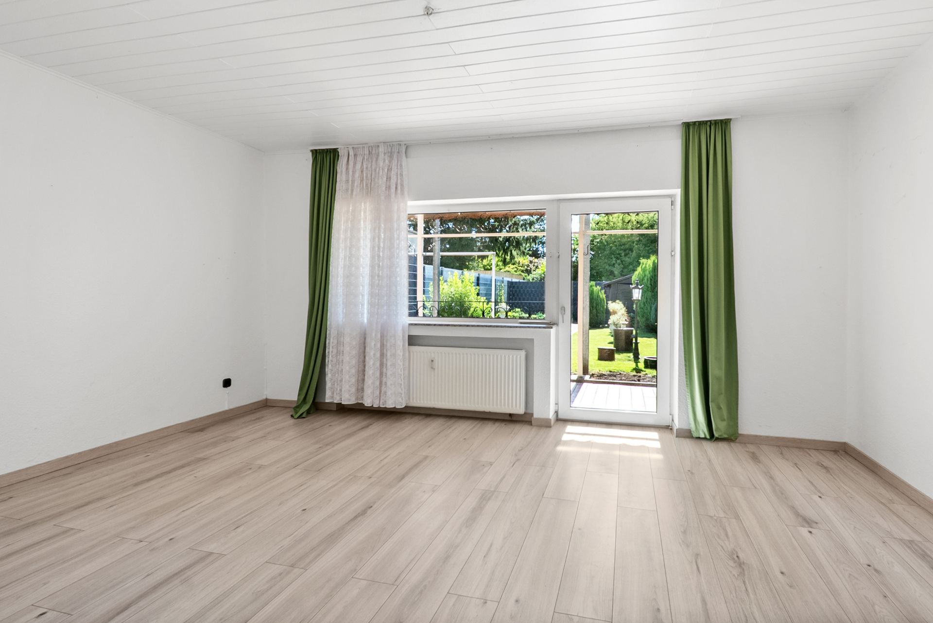 Heller leerer Raum mit Holzfußboden, Fenster, Tür zum Garten und grünen Vorhängen.