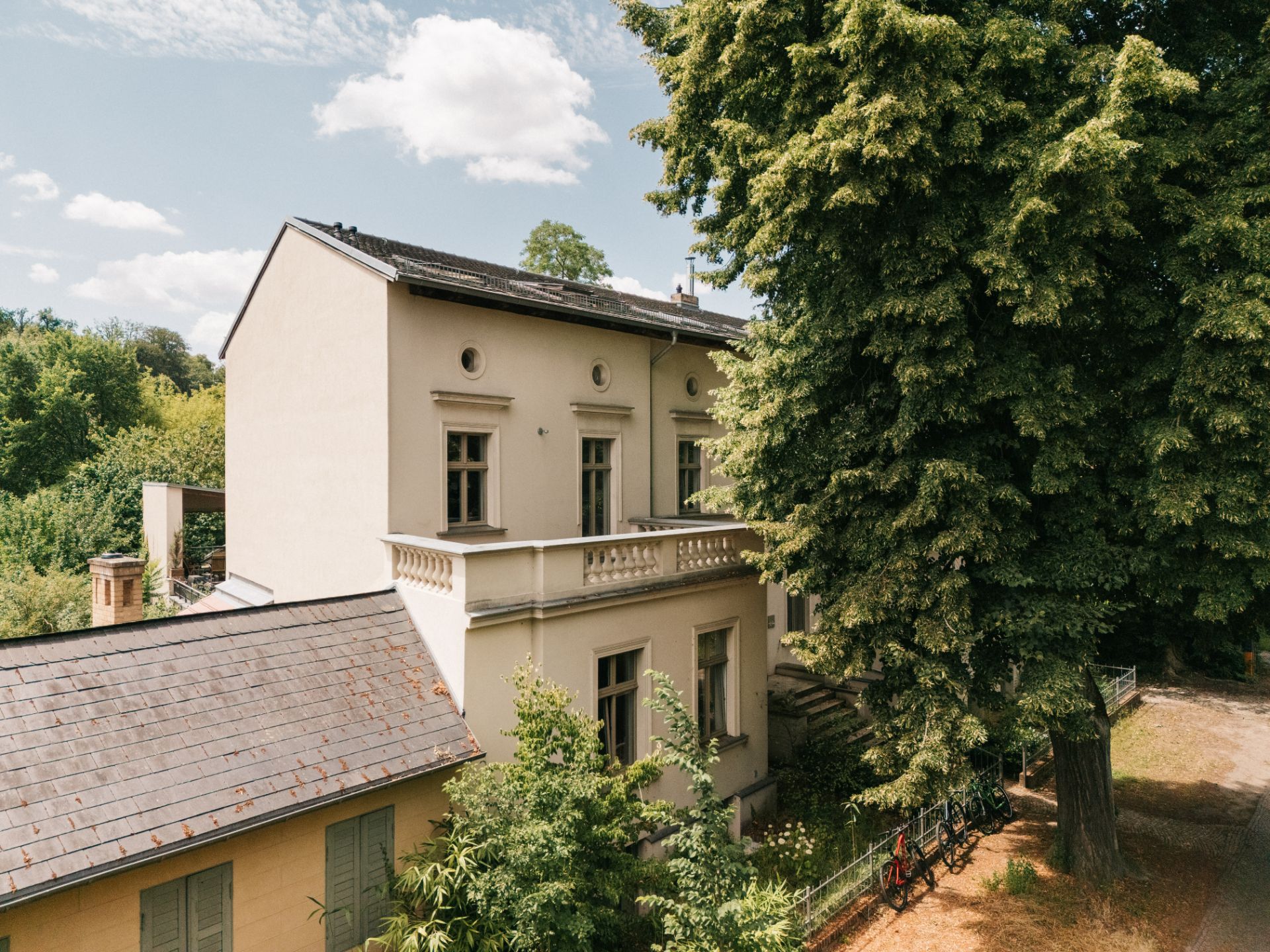 Außenansicht eines großen Wohnhauses mit Balkon, Bäumen und Garten im Vordergrund.