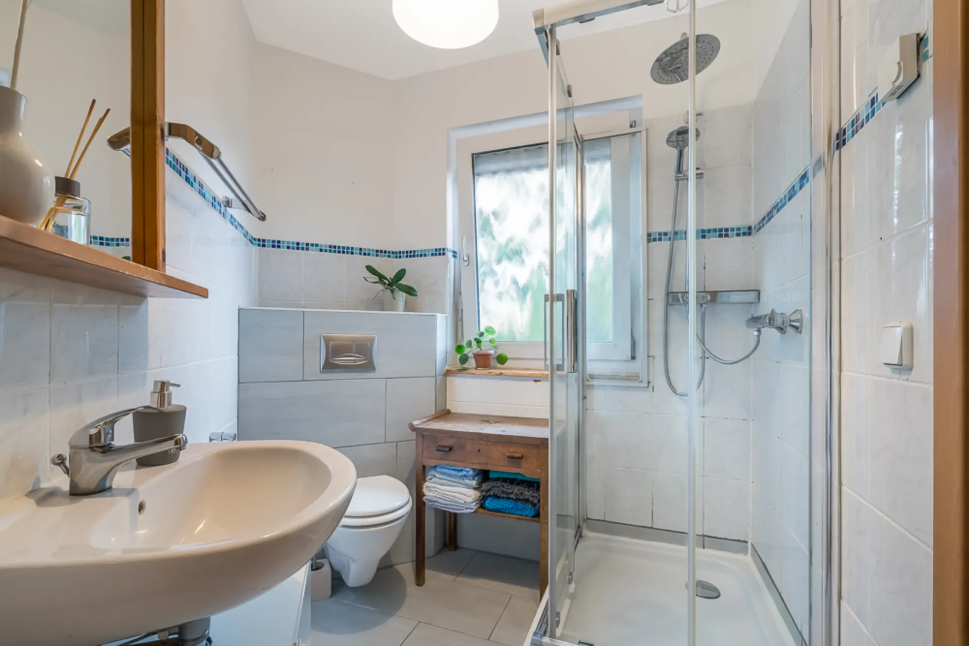 Modernes Badezimmer mit Dusche, Waschbecken, Toilette, Fenster und kleinem Holztisch mit Handtüchern.