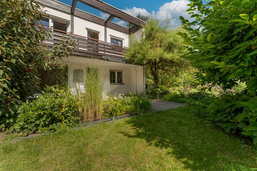 Zweistöckiges Wohnhaus mit Balkon und Garten, umgeben von vielen grünen Pflanzen.