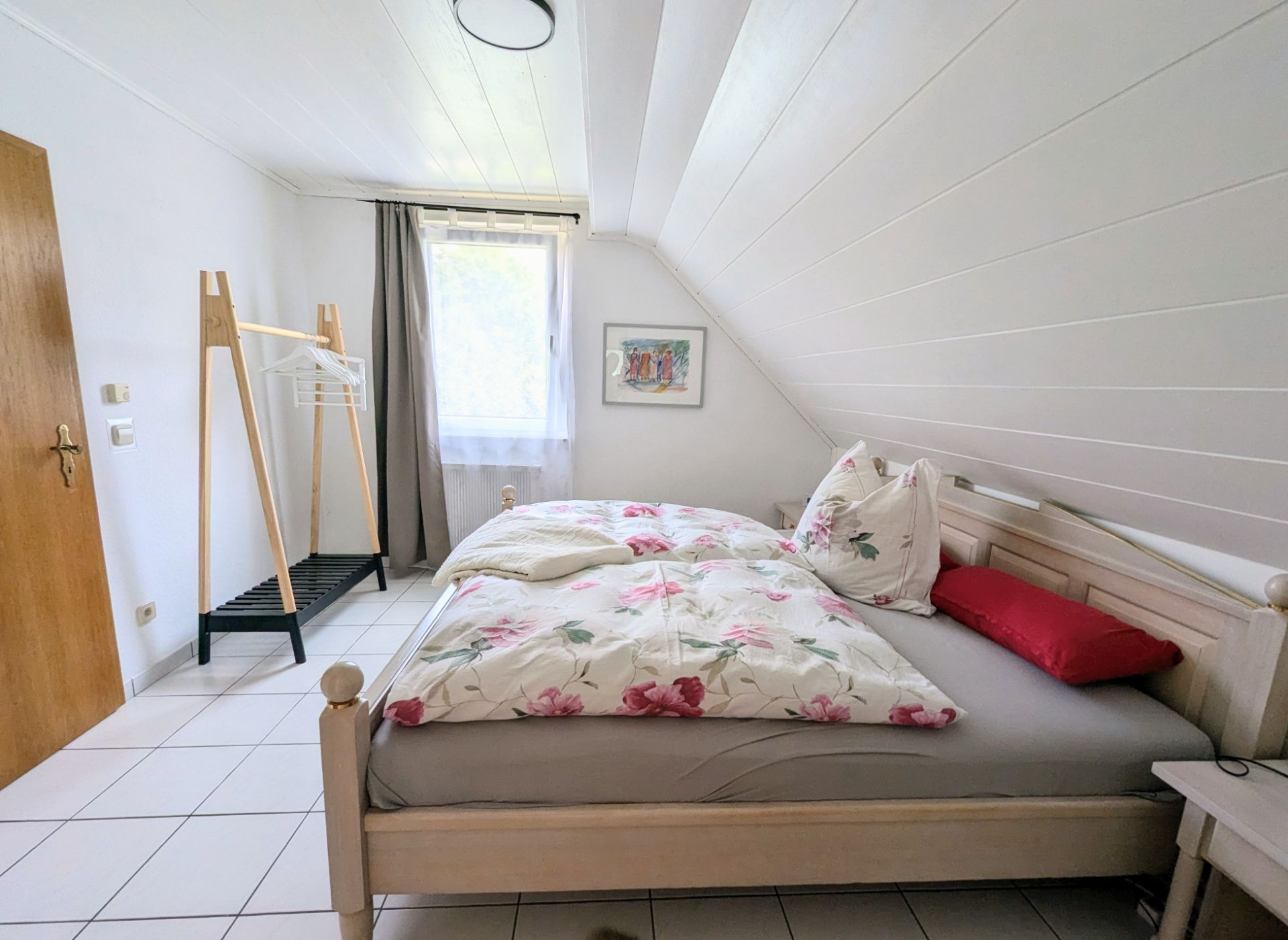 Helles Schlafzimmer mit Doppelbett, Kleiderständer und Fenster mit Vorhang.
