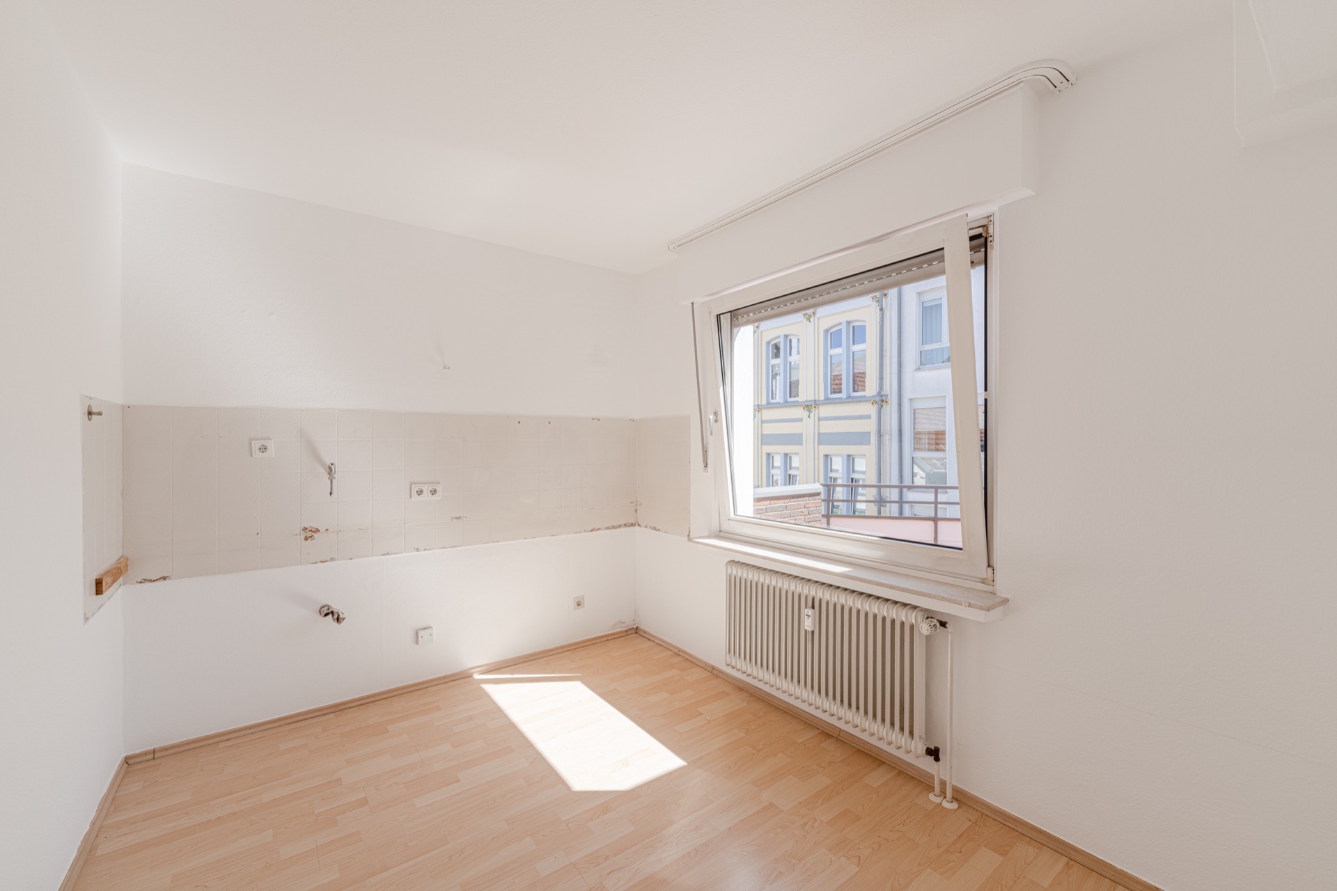 Leerer, heller Raum mit Laminatboden, offenem Fenster und Heizkörper unter dem Fenster.