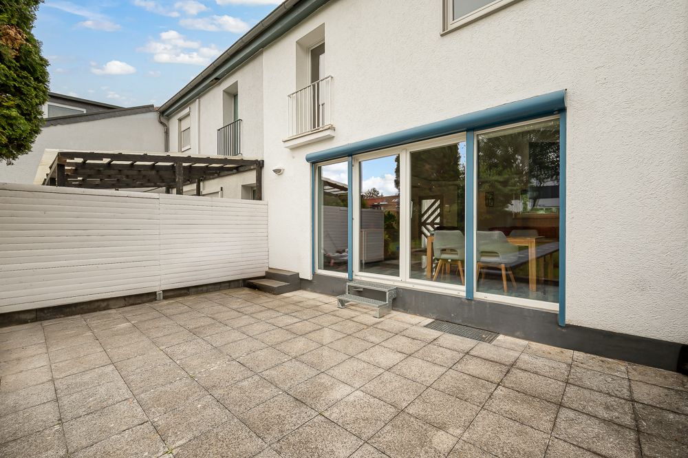 Gepflasterte Terrasse mit Sichtschutz an einem weißen Wohnhaus, große Fenster mit Blick ins Esszimmer.