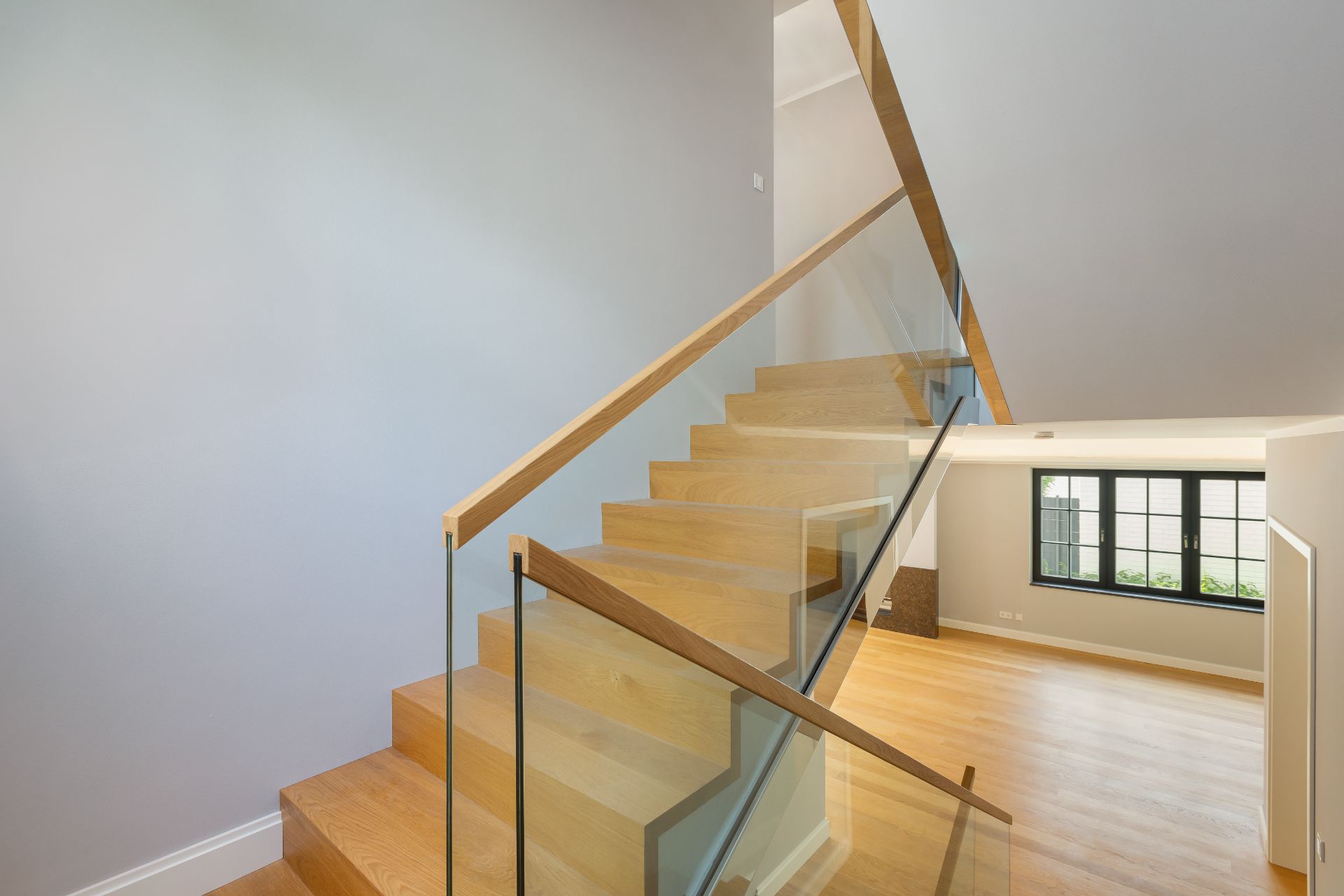 Holztreppe mit Glasgeländer in einem hellen Wohnraum mit Fenster.