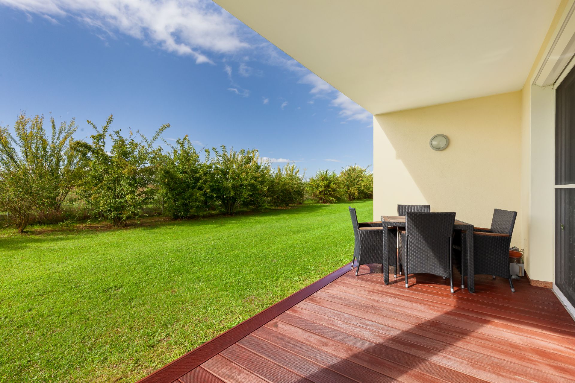 Terrasse mit Holzboden, Gartentisch und Stühlen, Blick auf einen großen grünen Garten mit Bäumen.